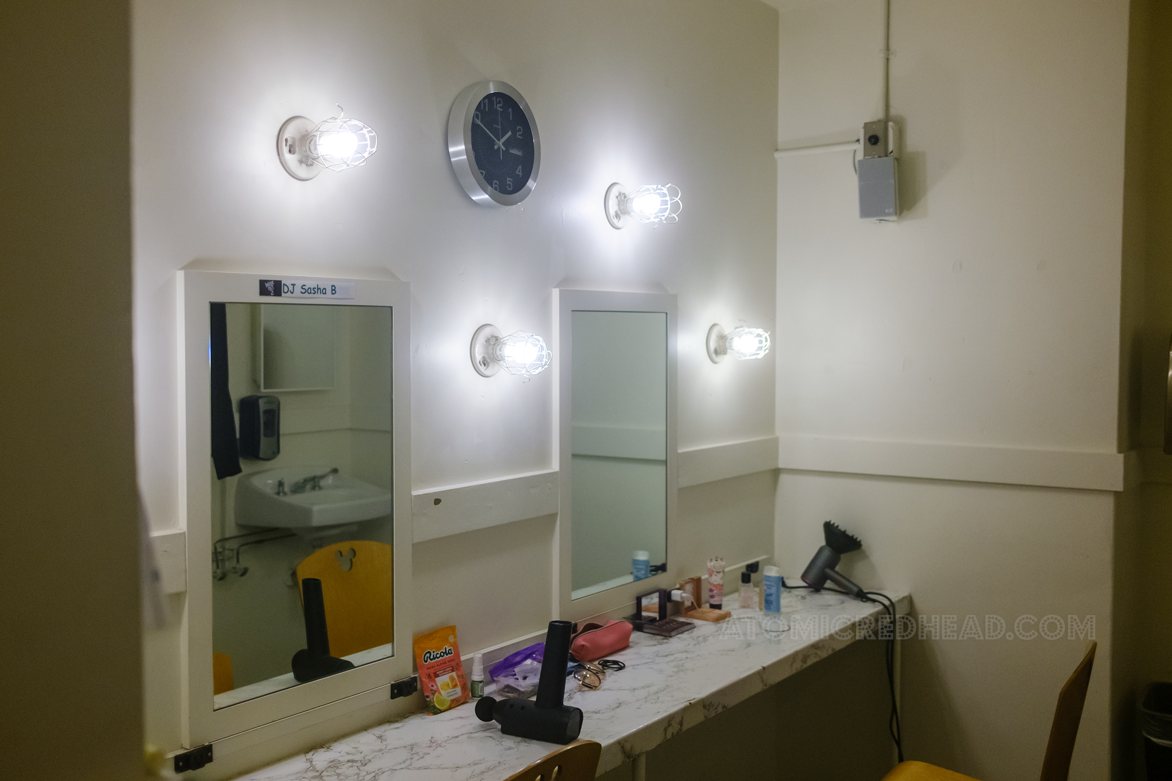 Inside a small dressing room, painted white with mirrors above a counter and lights on the wall.
