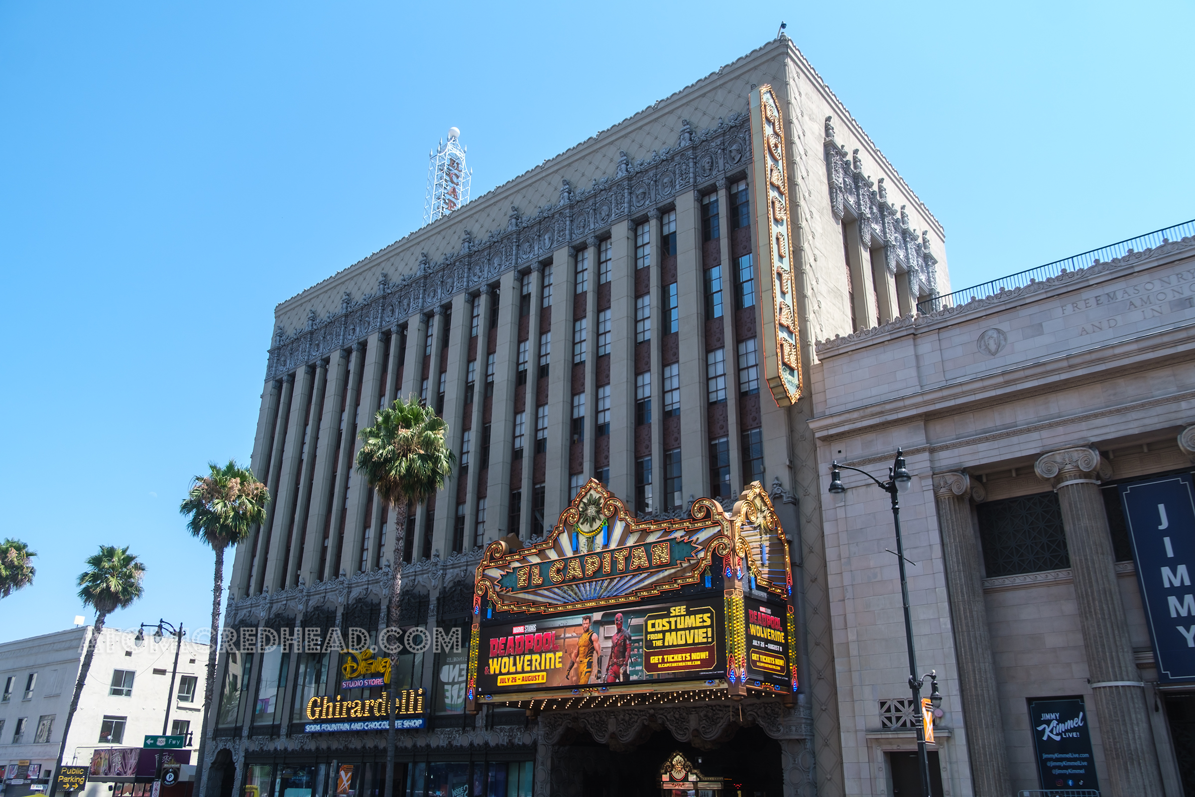 Overall exterior of the El Capitan, a multistory building with palm trees, a large lighted marquee reading "The El Capitan Theatre" in neon and bulb lights, and advertising Deadpool and Wolverine. Above a double sided, blade style neon and bulb sign reads "El Capitan" to the left in neon and bulb lights reads "Disney Studio Store Ghirardelli Soda Fountain and Chocolate Shop"