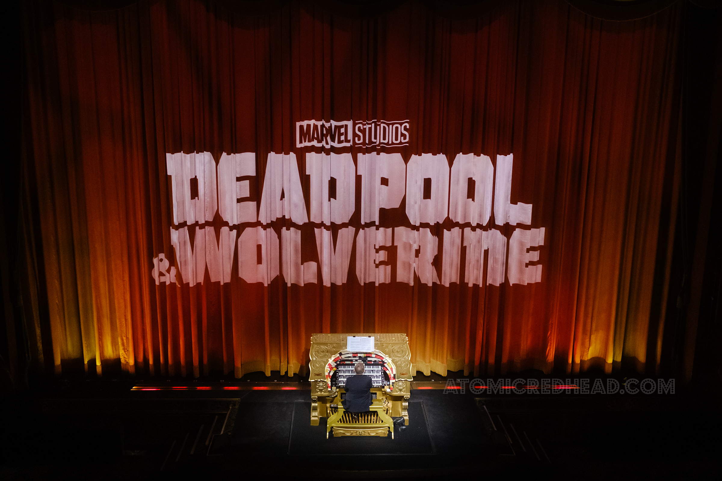Projected on a closed curtain reads "Marvel Studios Deadpool & Wolverine" and in front an organ player sits at the large gold organ.
