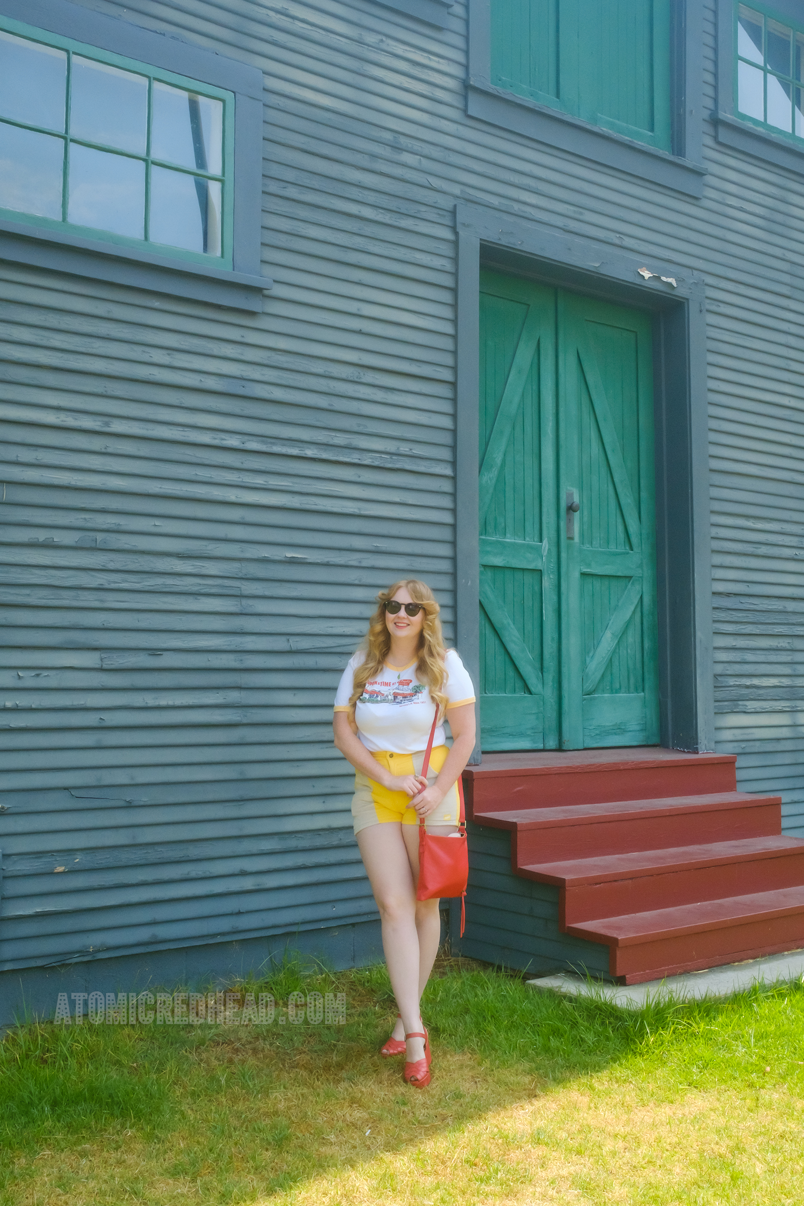 Myself, with my red hair down and wavy, wearing a white t-shirt featuring the exterior of the Mexican restaurant El Coyote on it, red and tan corduroy shorts, and a red purse and red shoes, standing outside the barn.