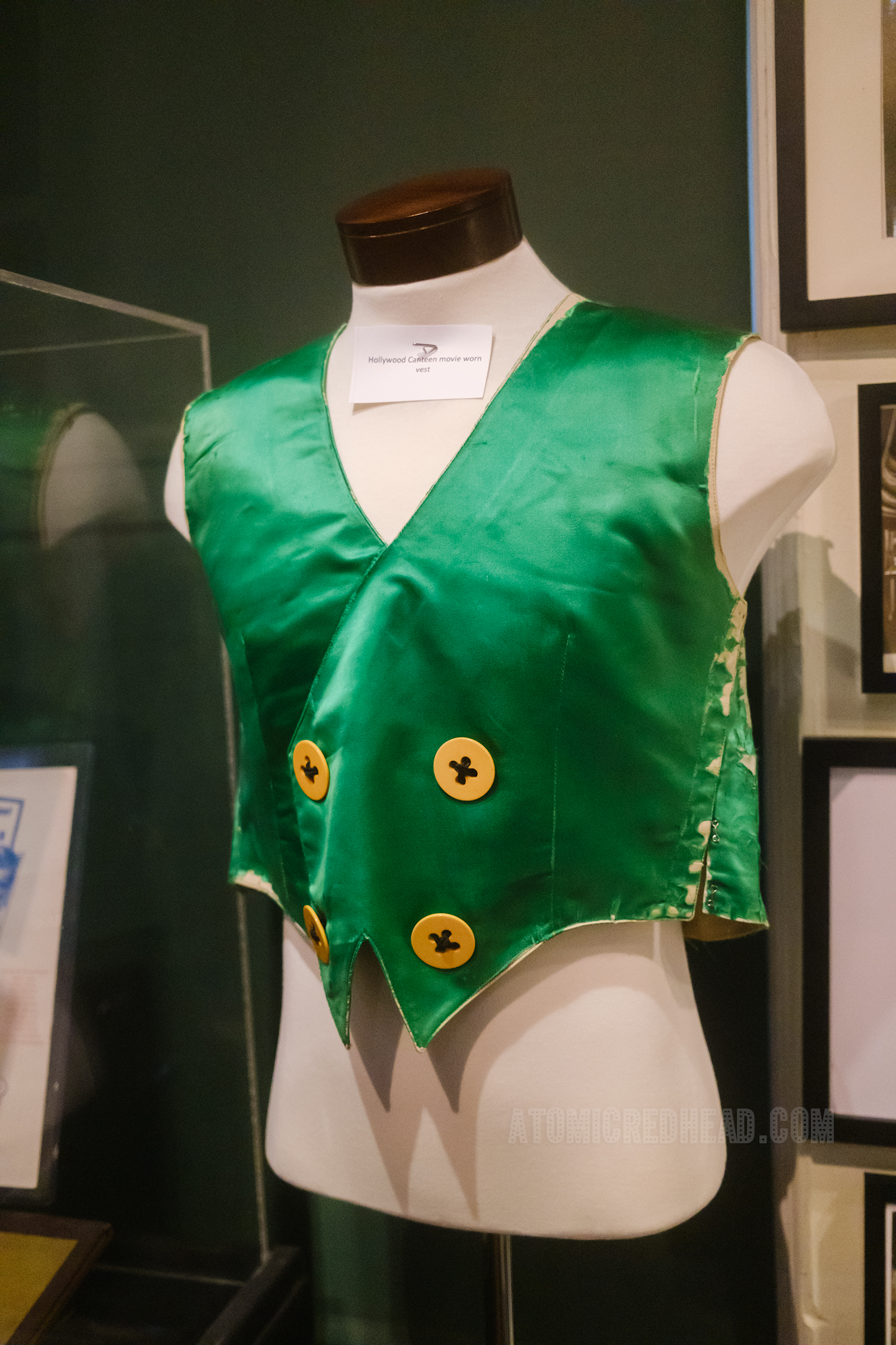 A small green satin vest with four large yellow buttons hangs on a white fabric covered torso.