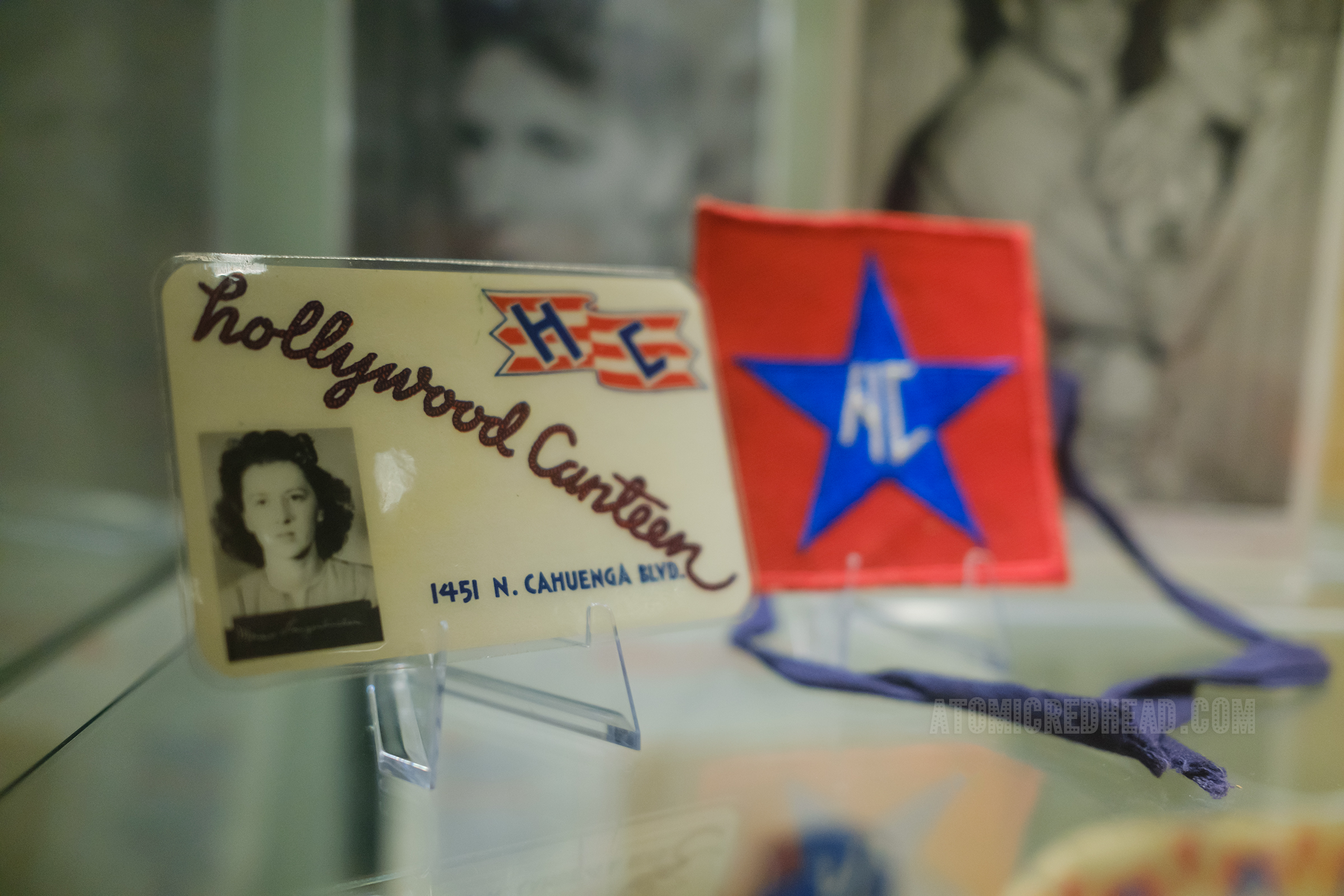 A laminated ID card of a dark haired woman, her photo sits in the lower left. Diagonally across the white card in rope script reads "Hollywood Canteen" and in the upper right corner is the logo, a red and white stripe waving flag with the letters "HC" in blue. Behind the card sits a red square armband that features a large blue star and the letters "HC" embroidered in white.