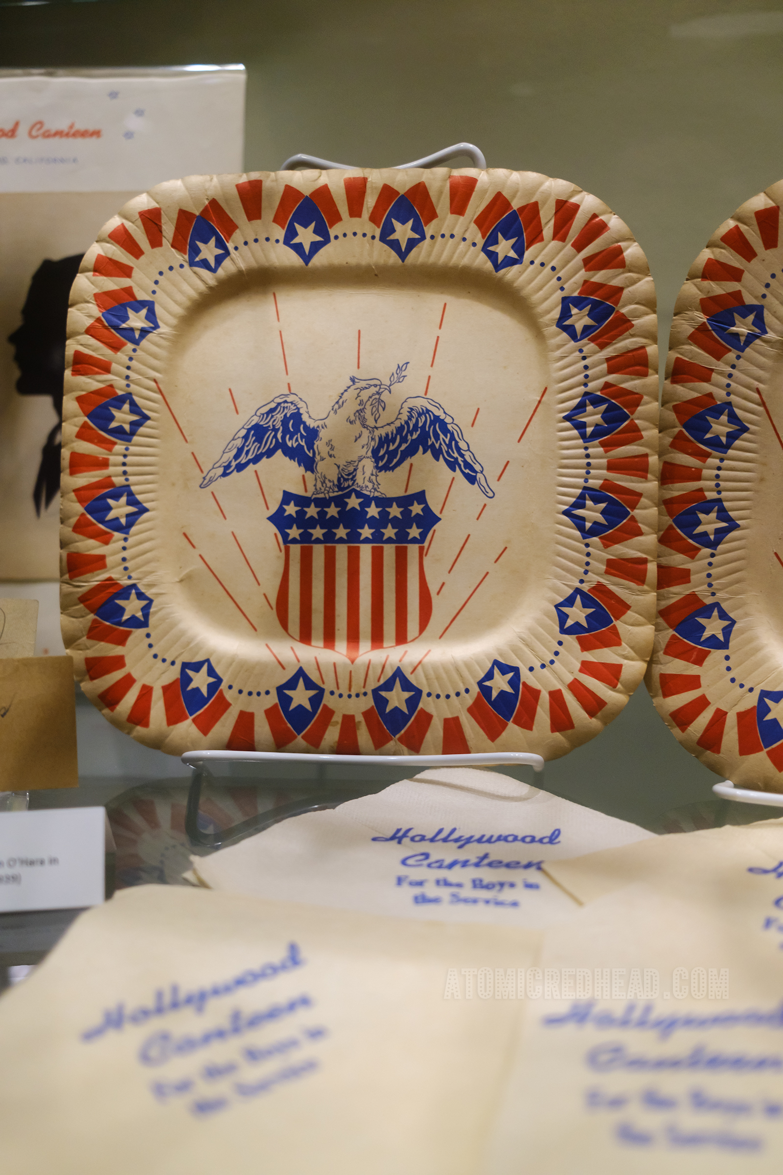 A paper plate with a patriotic motif of a red and white stripe, and blue and white star shield and an eagle. Below are napkins, that are white with blue lettering reading "Hollywood Canteen For the Boys in the Service"