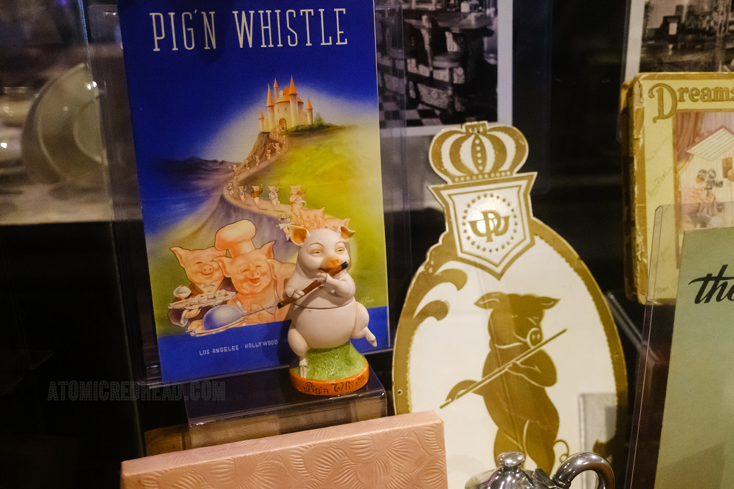 Artifacts from Pig'n Whistle, including a menue featuring pigs dancing down a path, and a ceramic figure of a pig with a flute.