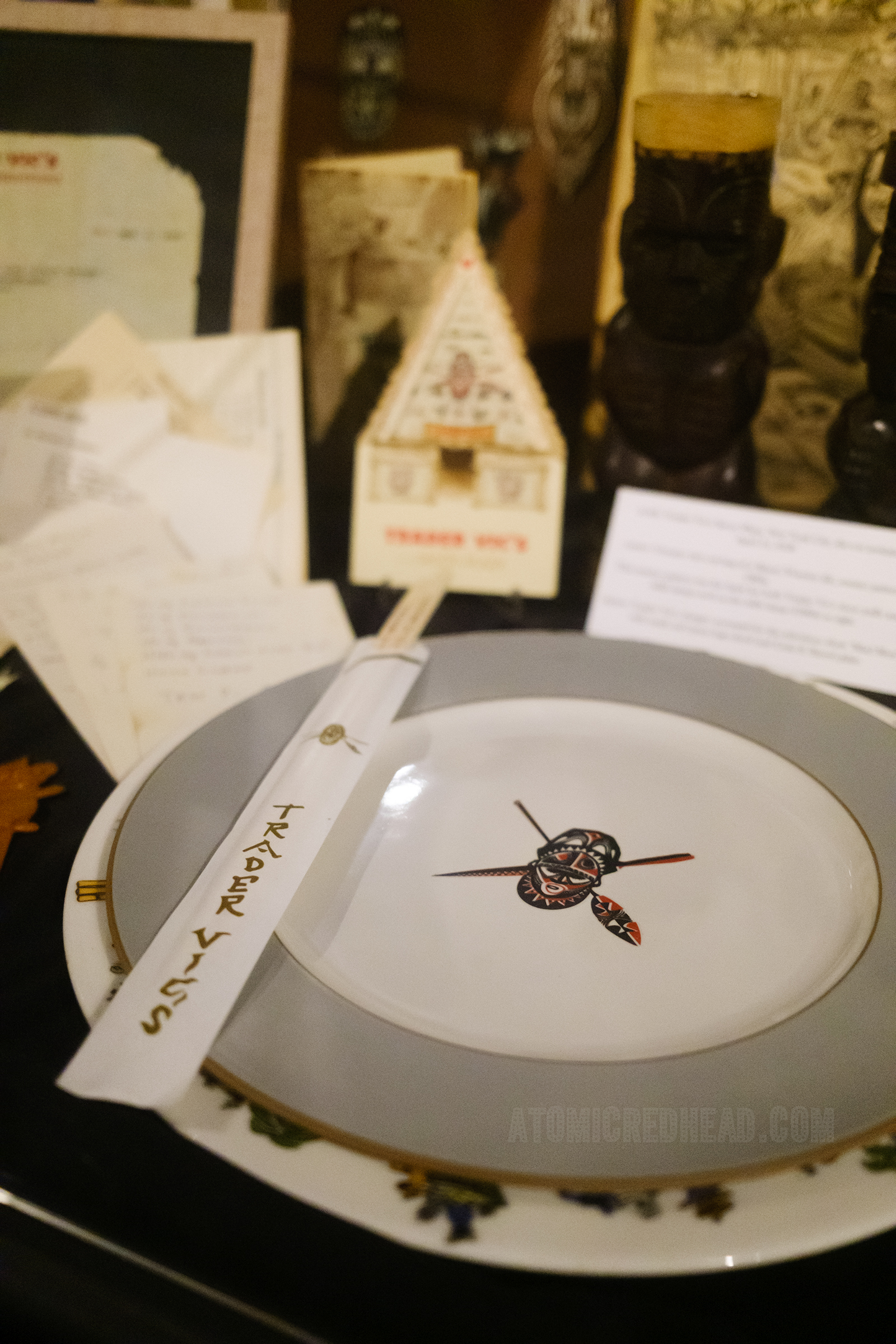 A grey and white plate features a tiki mask and crossed spears along with a set of chopsticks, the sleeve reads "Trader Vic's"