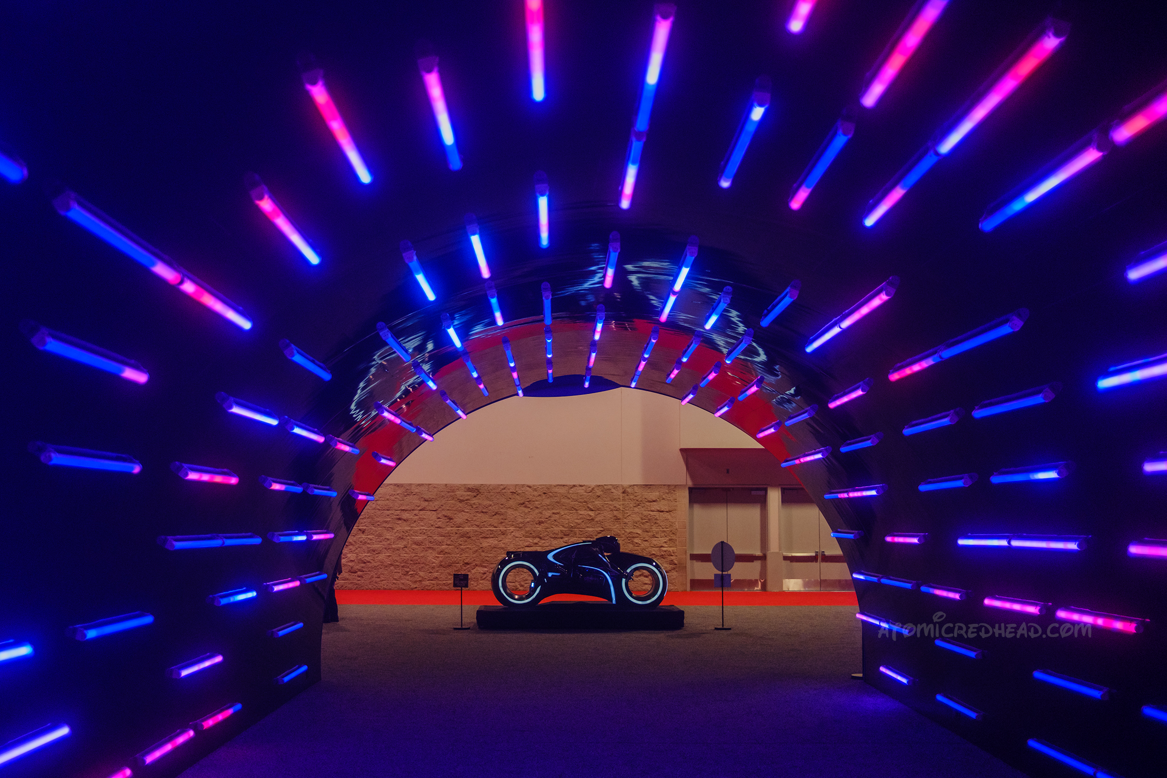 An arched tunnel features blue and pink lights ending in a futuristic motorcycle known as a Lightcycle from Tron: Legacy.