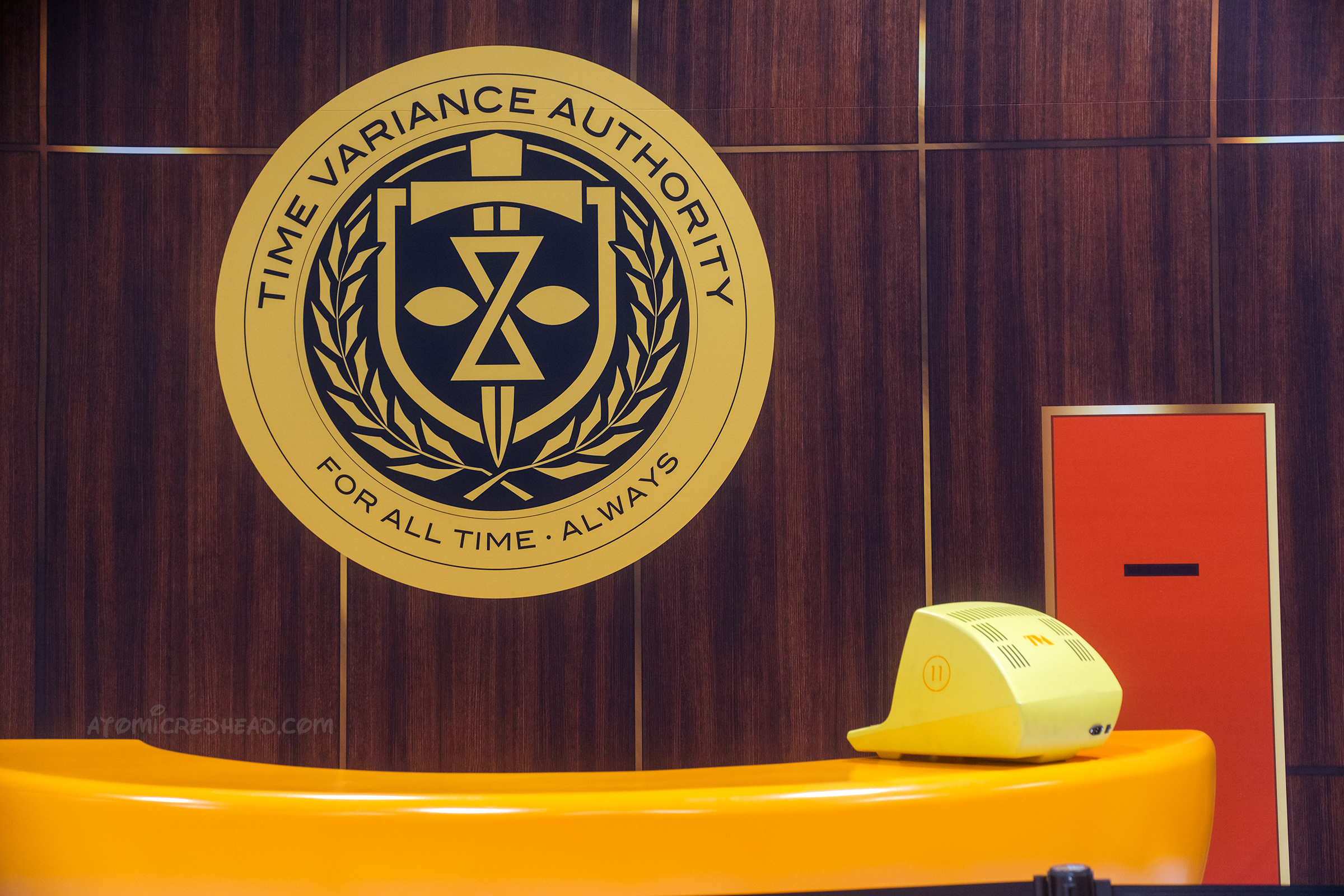 On a wood panel wall sits the seal of the TVA and text reading "Time Variance Authority For All Time Always" below is a curved yellow desk with a yellow computer on it.