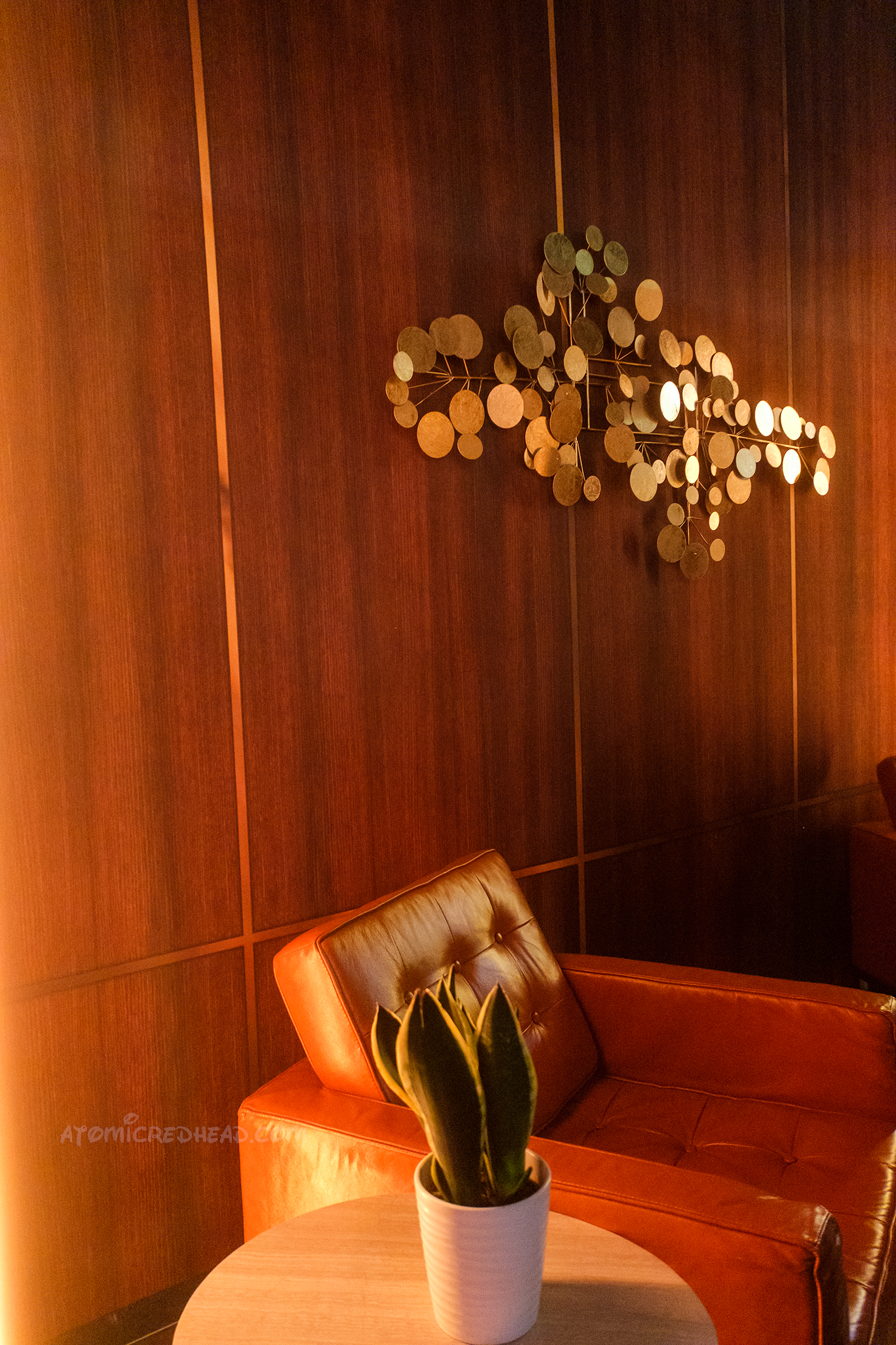 A gold sculpture of circles hangs on a wood panel wall, below is an orange chair.