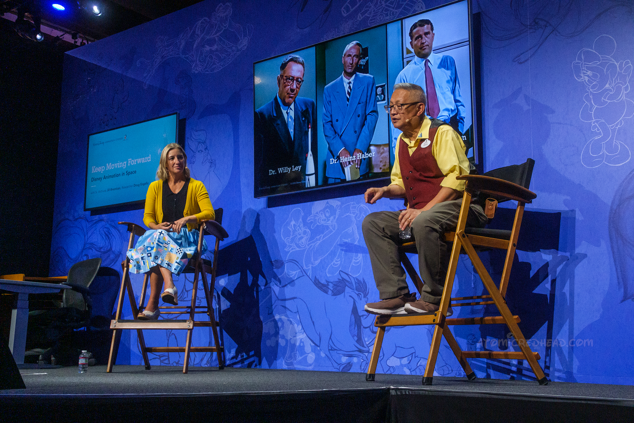 Two people sit on chairs with a screen behind them displaying images of rocket scientists on it.