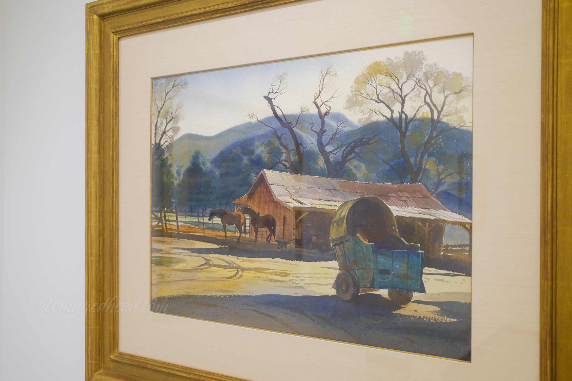A watercolor of two horses stand in front of a small barn, in the foreground is a cart.