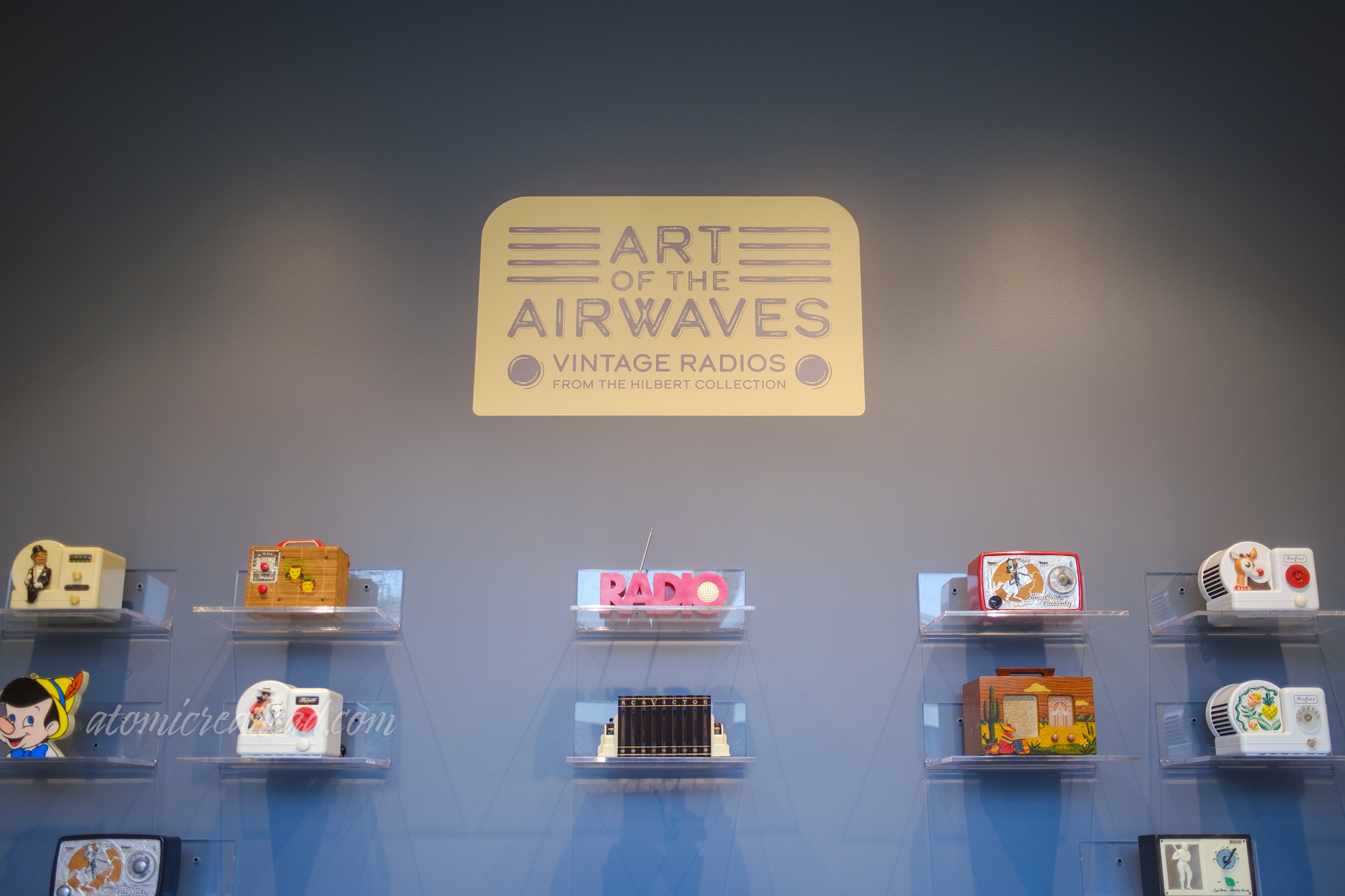 Various radios sit on shelves, text at the top reads "Art of the Airwaves Vintage Radios from the Hilbert Collection"