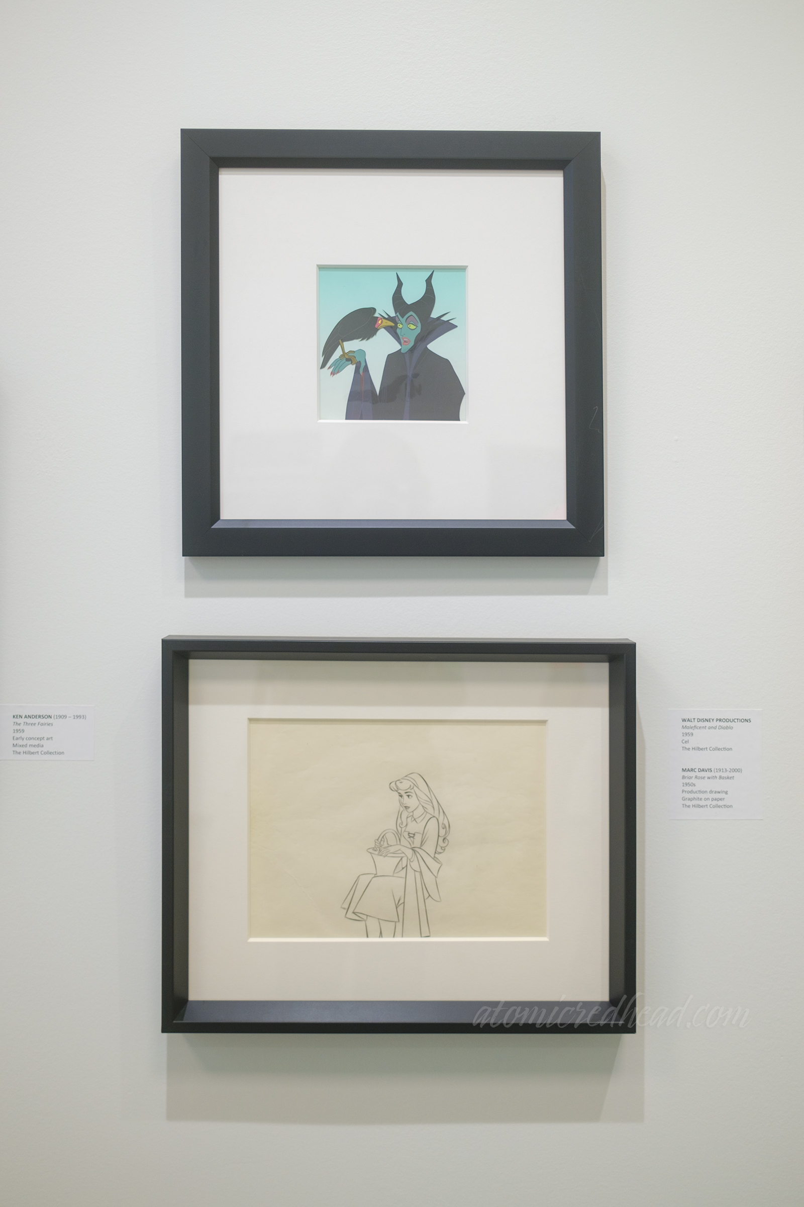 Two framed pieces, the top an animation cel of Maleficent, with her green skin and crow companion on her hand, below a production drawing of Briar Rose with a basket.