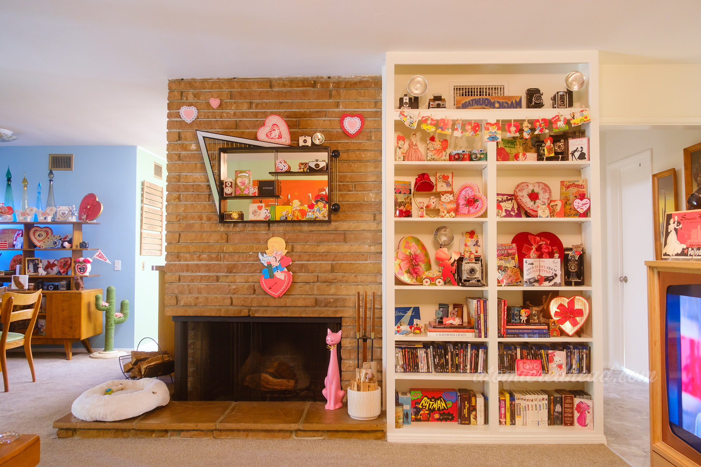On the left is our fireplace, with a shadow box hanging above, inside are various vintage valentines. To the right is a built in bookcase with decor on it.