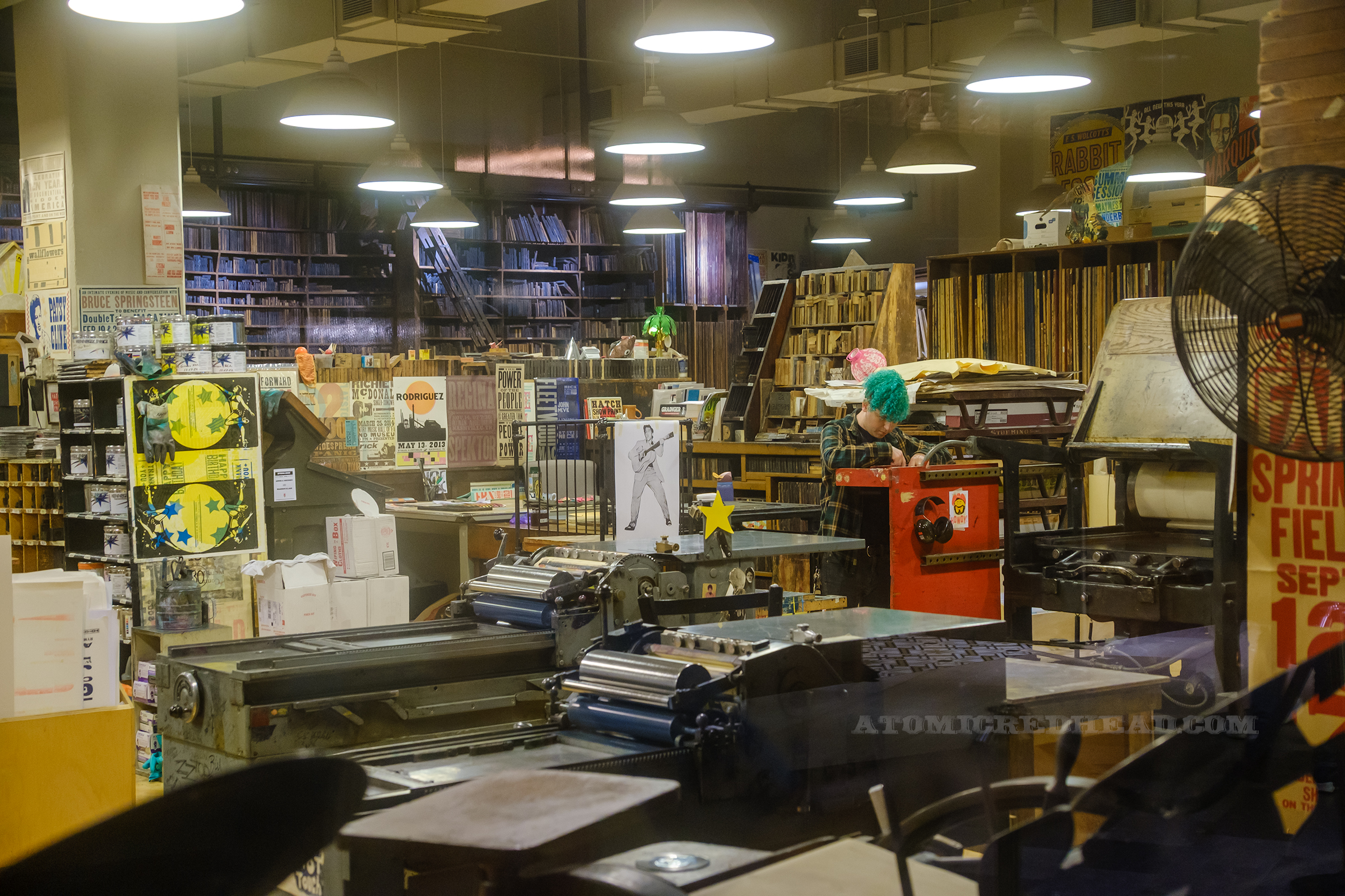 View of the workshop, along the back walls are shelves full of blocks, other areas have desks, shelves, and various prints scattered and hung, a large printing press sits closer to the window.