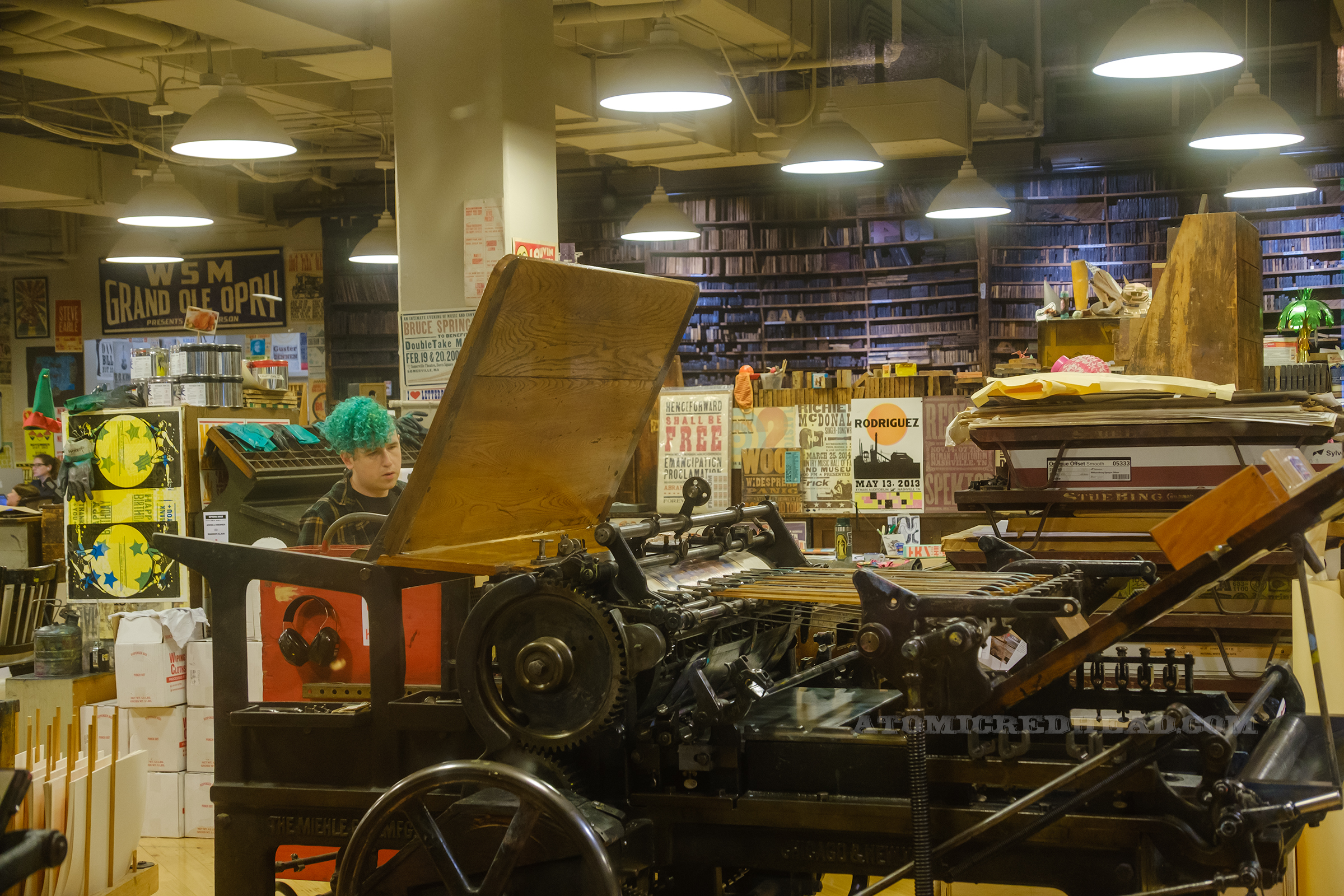 View of the workshop, along the back walls are shelves full of blocks, other areas have desks, shelves, and various prints scattered and hung, a large printing press sits closer to the window.