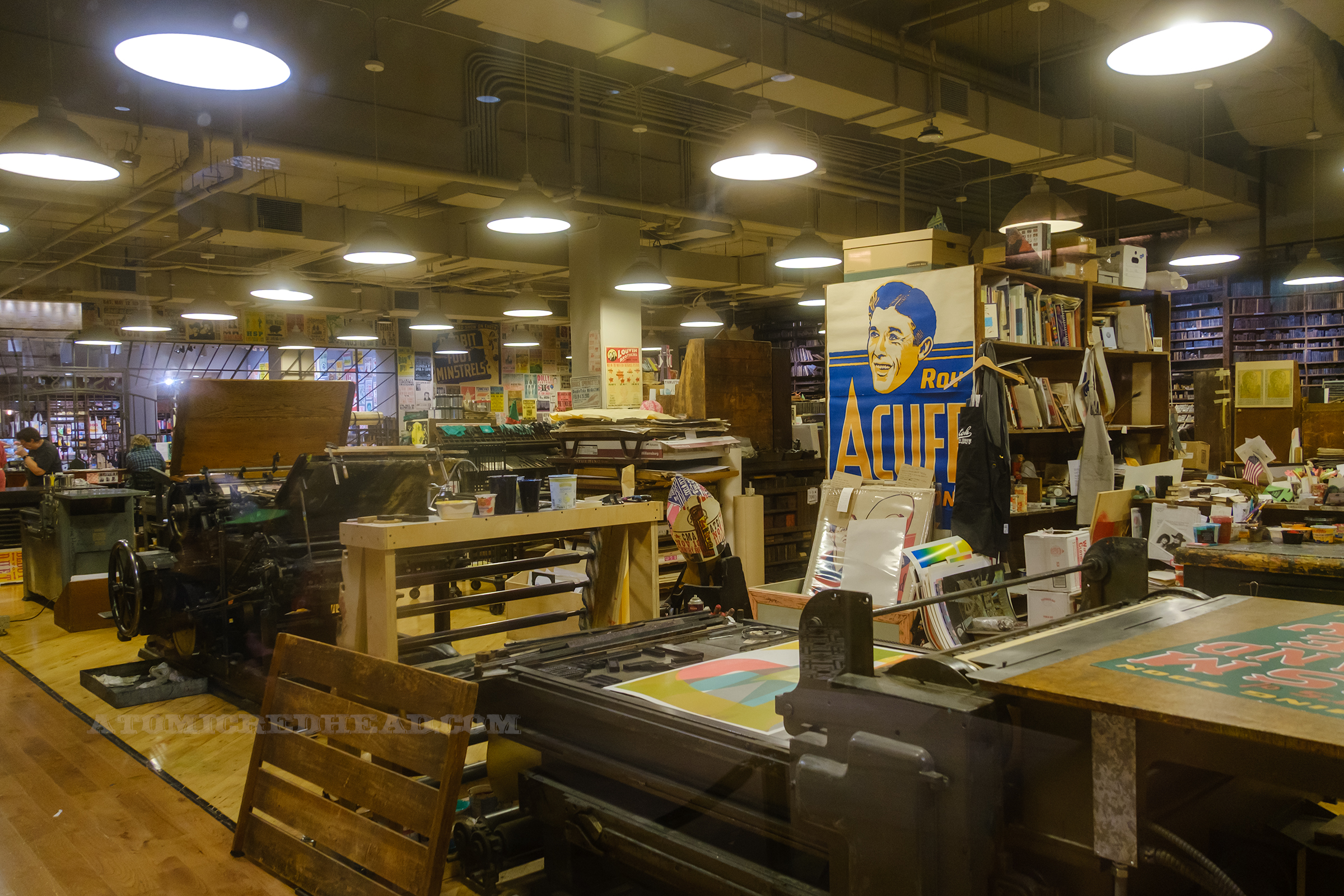 View of the workshop, along the back walls are shelves full of blocks, other areas have desks, shelves, and various prints scattered and hung, a large printing press sits closer to the window.