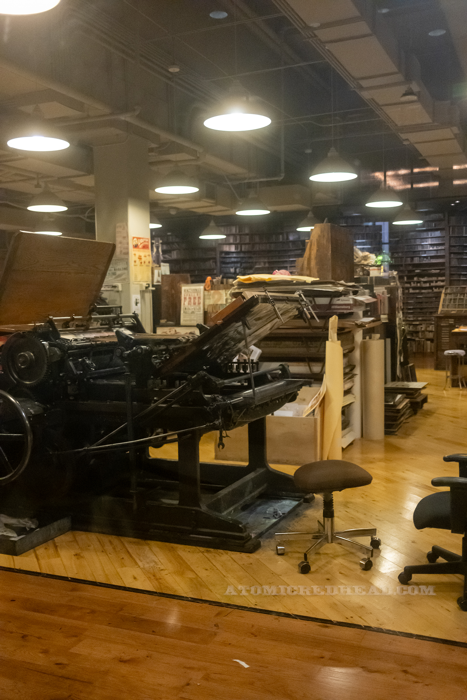 View of the workshop, along the back walls are shelves full of blocks, other areas have desks, shelves, and various prints scattered and hung, a large printing press sits closer to the window.