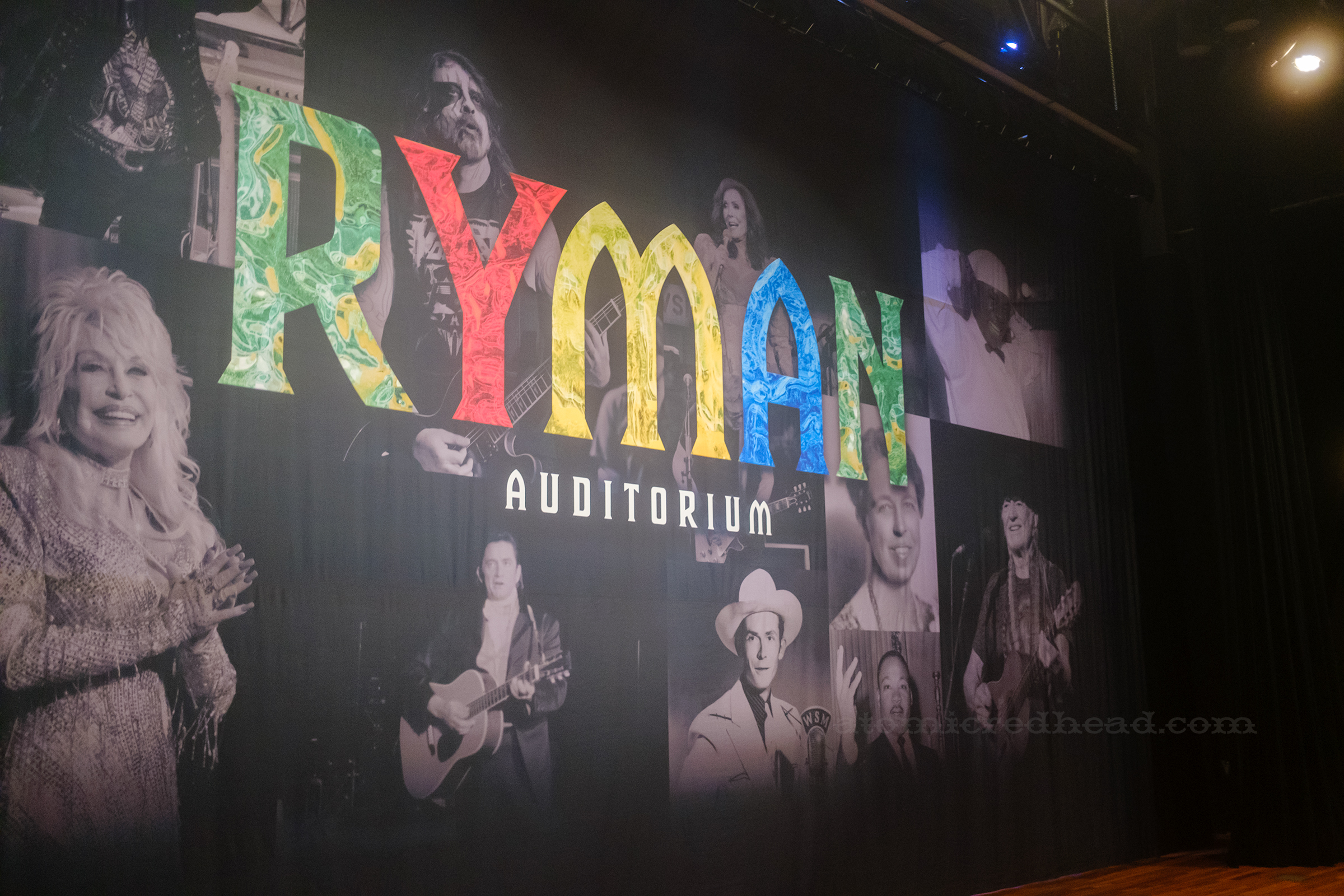 A large backdrop features black and white images of people who have performed on the Ryman stage, including Dolly Parton, Hank Williams, Johnny Cash, Willie Nelson, with large red, yellow, blue, and green letters reading "Ryman Auditorium"