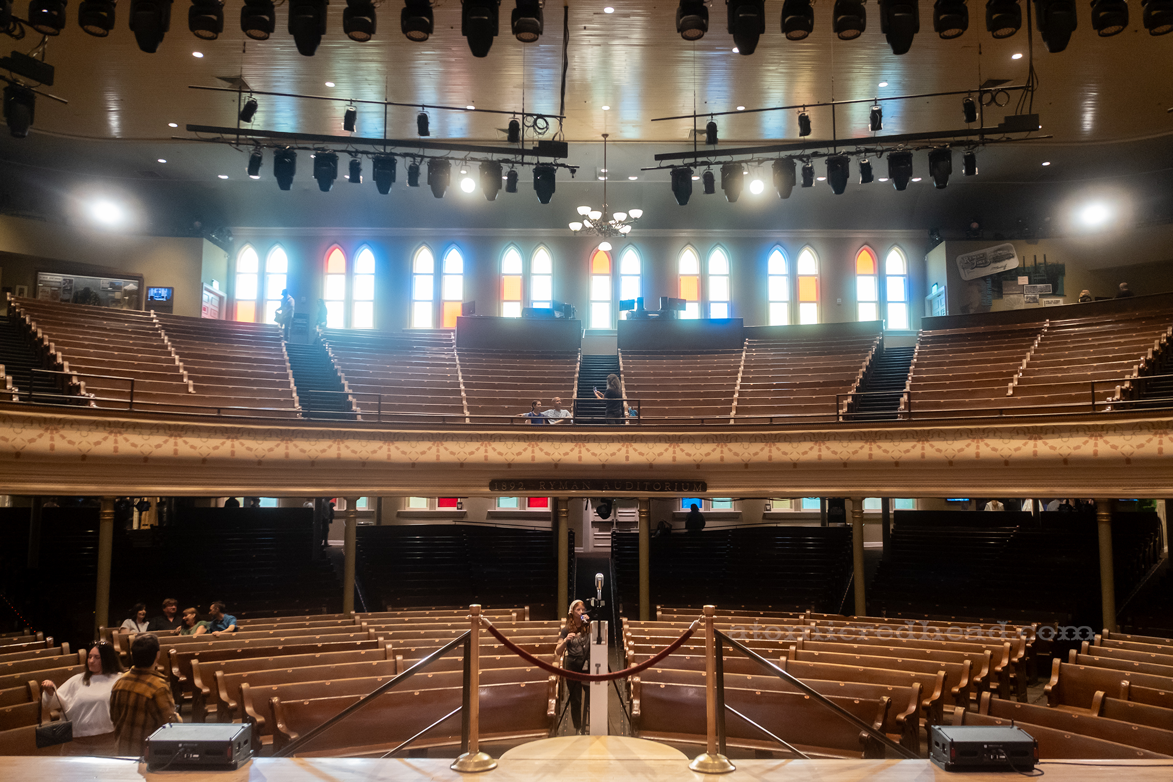 Overall image of the auditorium from the view of the stage. Rows of pews curve around the stage with tall stained glass windows of blue, yellow, red, and green along the back.