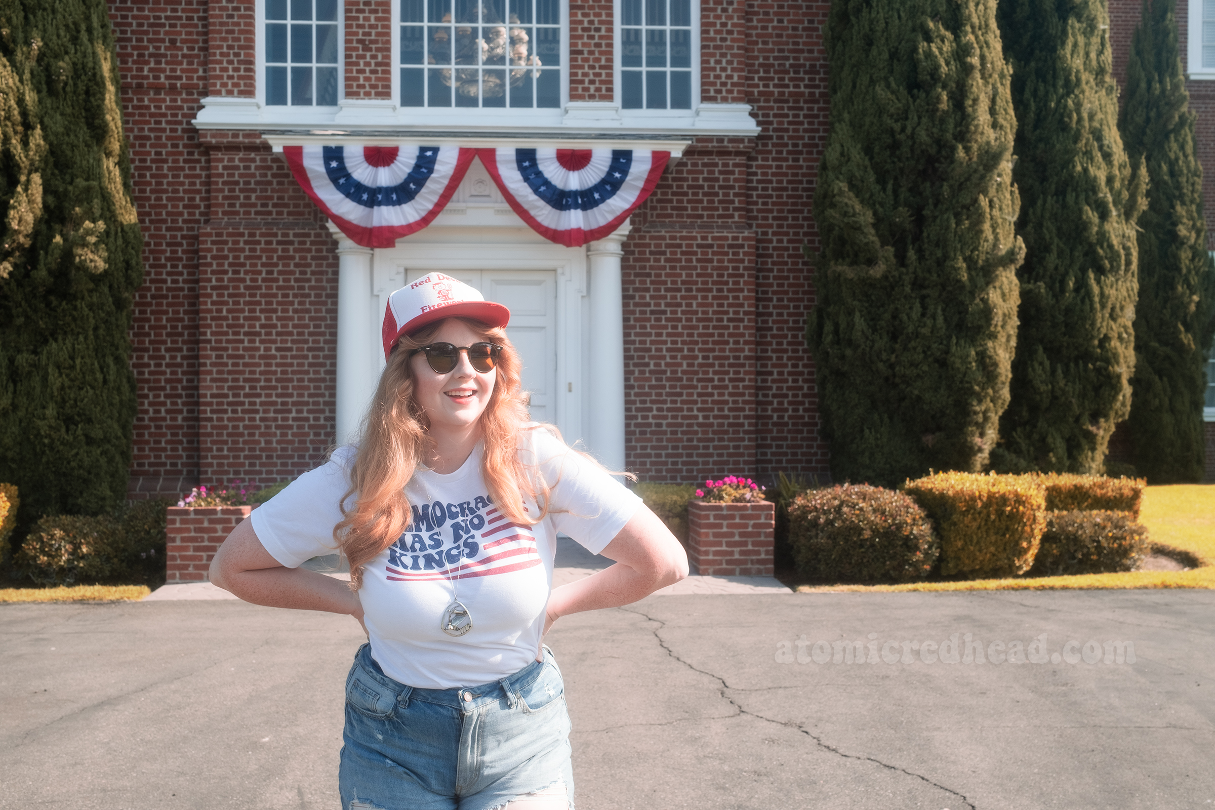 Myself, wearing a red and white hat that reads "Red Devil Fireworks" a white shirt with a stylized American flag that reads "Democracy Has No Kings" and blue jean shorts, standing in front of Knott's Berry Farm's recreation of Independence Hall.