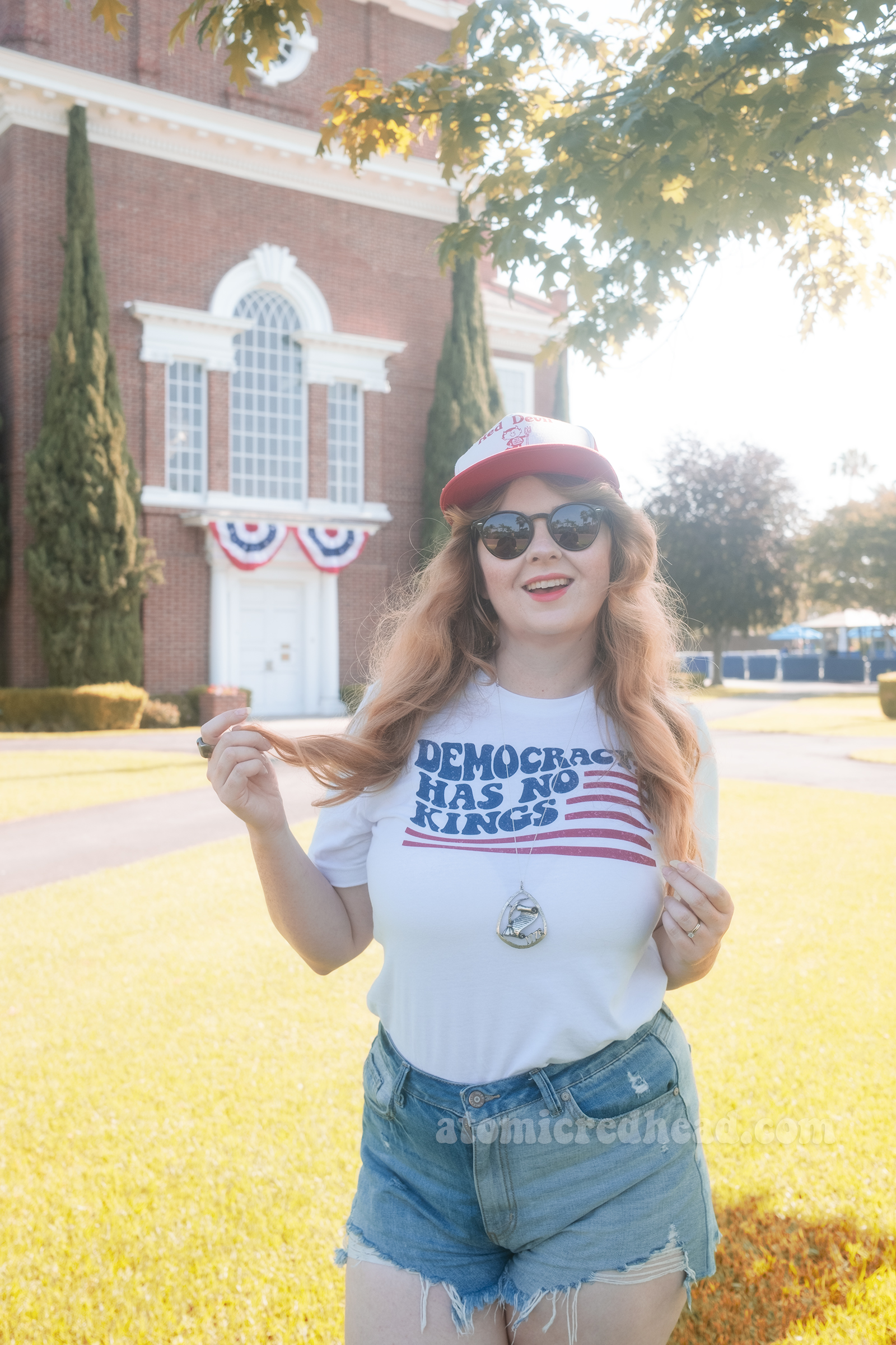 Myself, wearing a red and white hat that reads "Red Devil Fireworks" a white shirt with a stylized American flag that reads "Democracy Has No Kings" and blue jean shorts, standing in front of Knott's Berry Farm's recreation of Independence Hall.