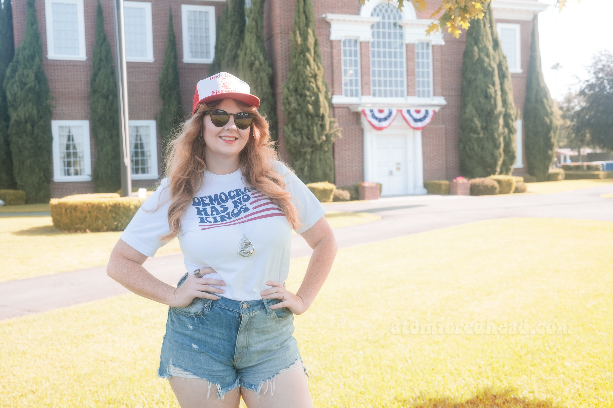 Myself, wearing a red and white hat that reads "Red Devil Fireworks" a white shirt with a stylized American flag that reads "Democracy Has No Kings" and blue jean shorts, standing in front of Knott's Berry Farm's recreation of Independence Hall.