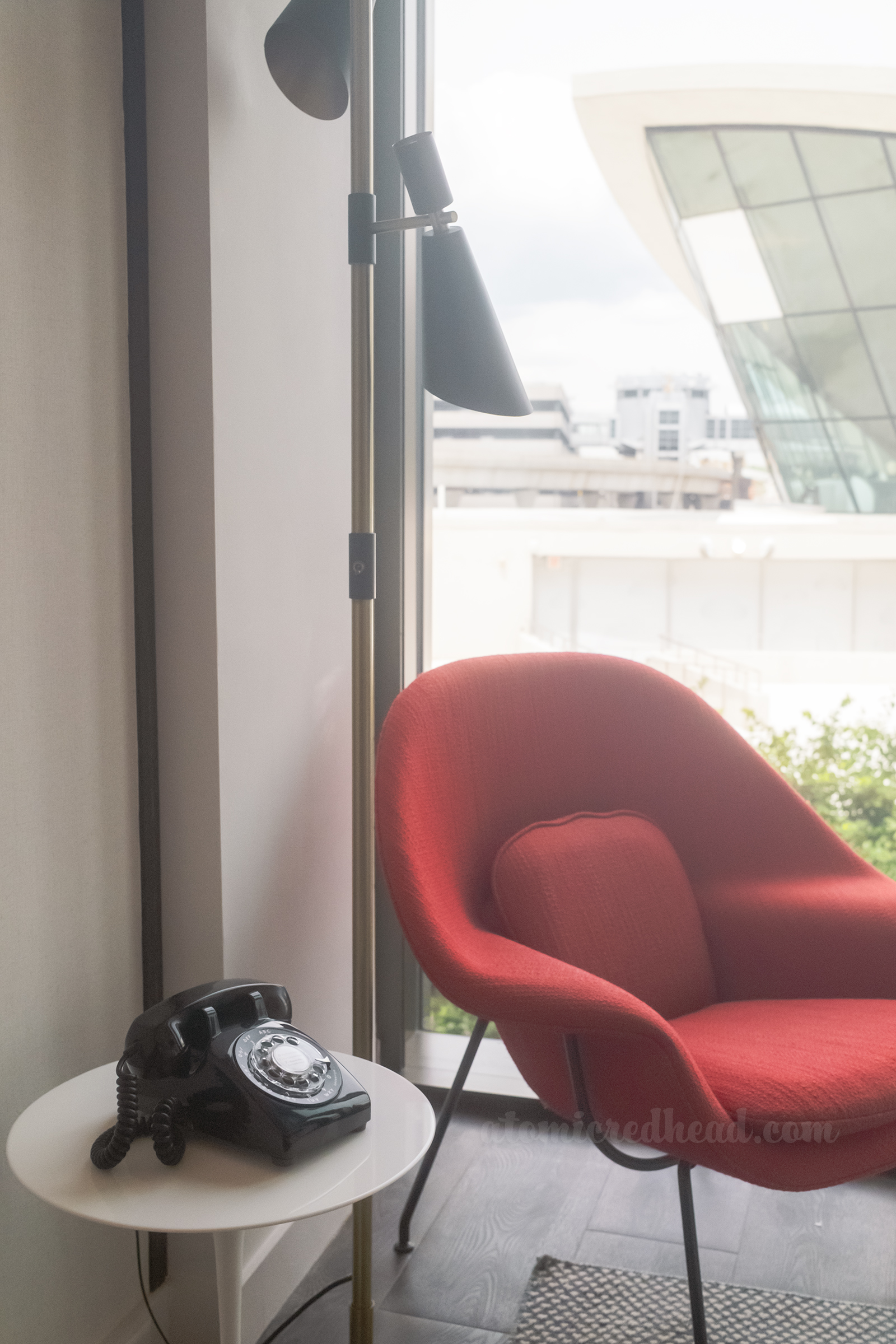 The red "Womb" chair in the room sits next to a window and a white table with a black phone on it.