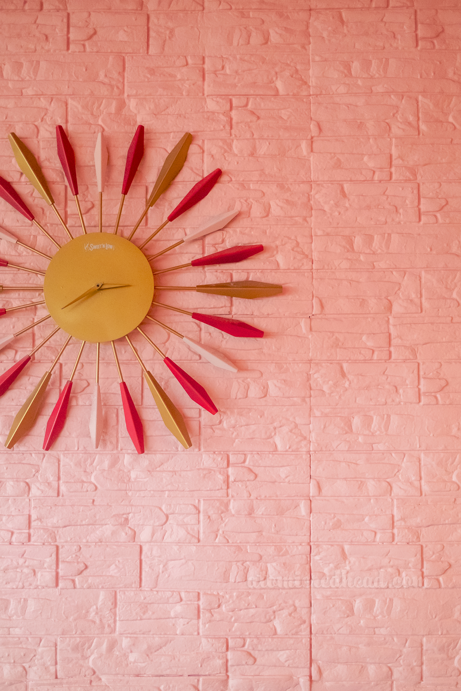 Close-up of a pink and gold starburst clock hanging on a pink stone wall.