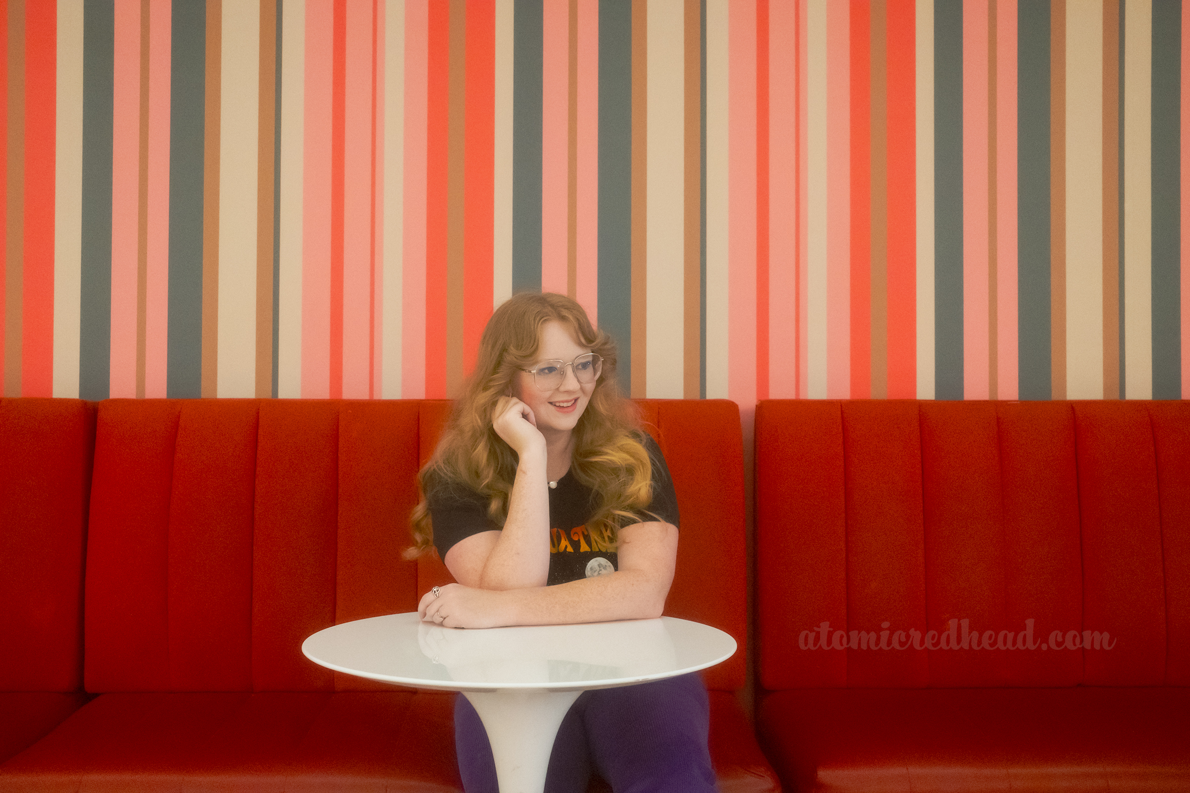Myself seated in on a red upholstered bench with pink, green, gold, and cream stripe wallpaper.