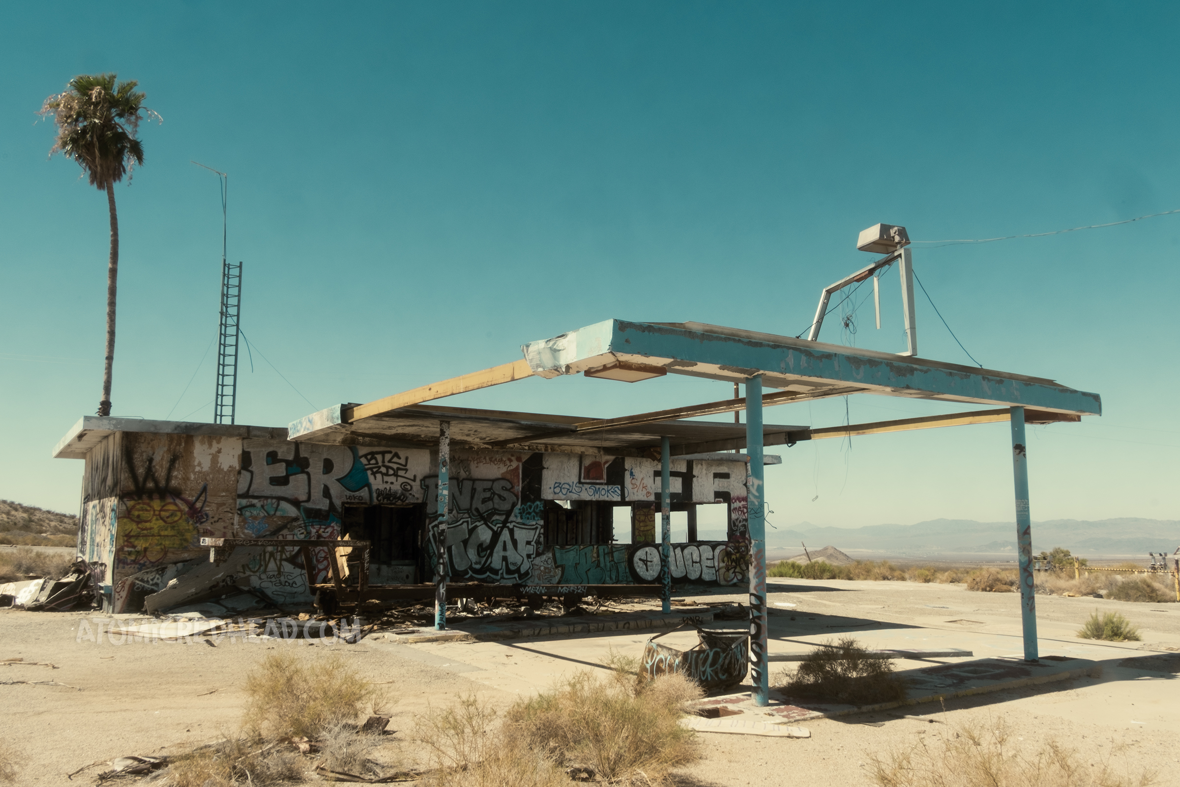 An abandoned gas station in the middle of the desert, a palm tree towers over it on the left side. The building has graffiti covering the majority of it.