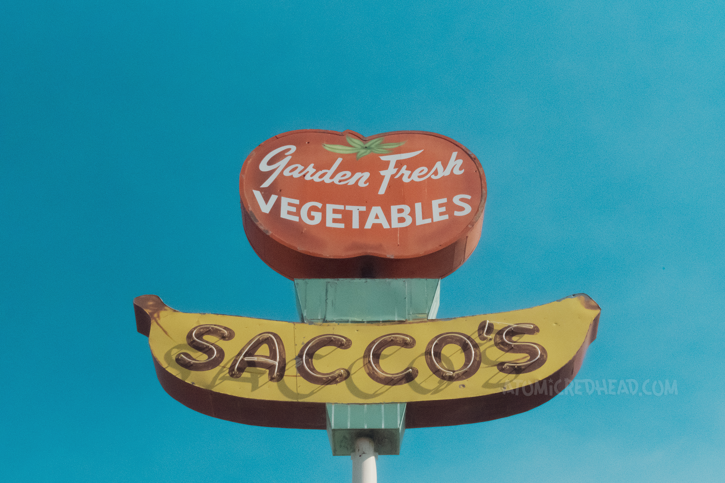 A large sign features a tomato at the top reading "Garden Fresh Vegetables" and below a banana with neon letters reading "Sacco's"