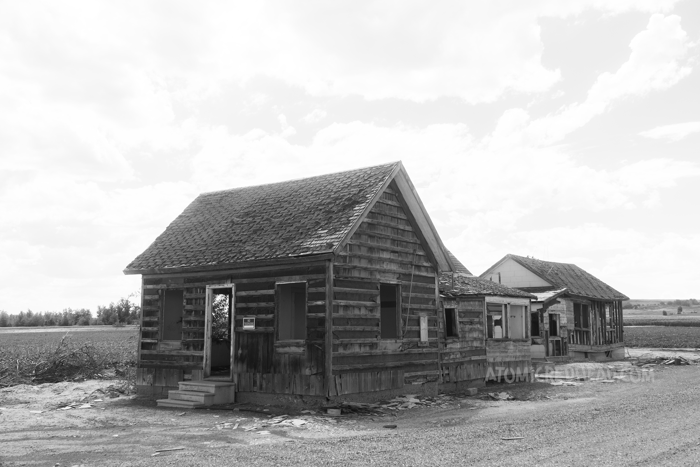 A black and white image of two abandoned buildings with open door and windows.