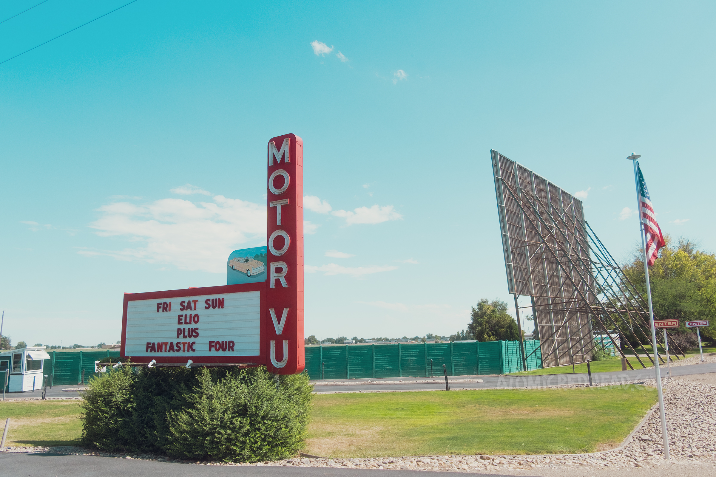 Overall of the Motor-Vu drive-in. A neon sign reads "Motor-Vu" on the left, with an image of a car, on the left is the towering screen.