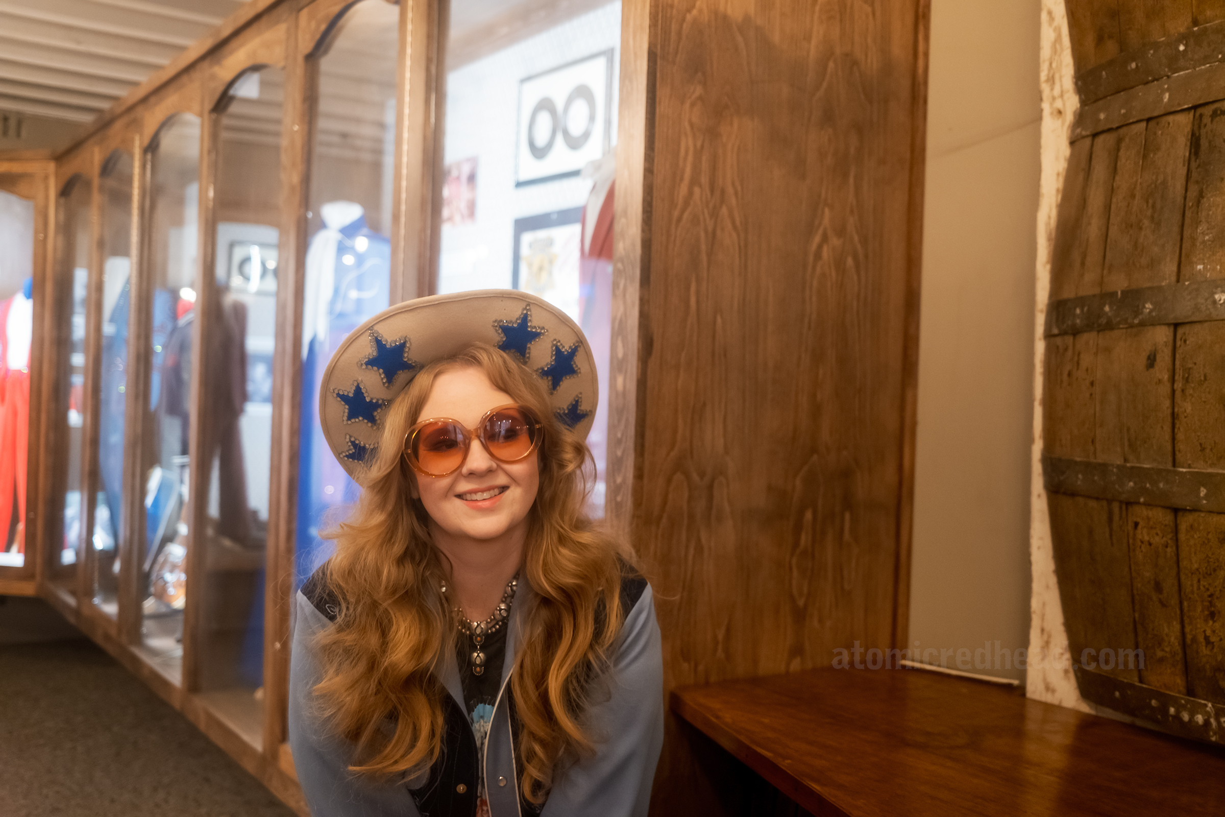 Myself, wearing a blue western wear shirt, cream cowboy hat with blue stars, leaning against a barrel bar.