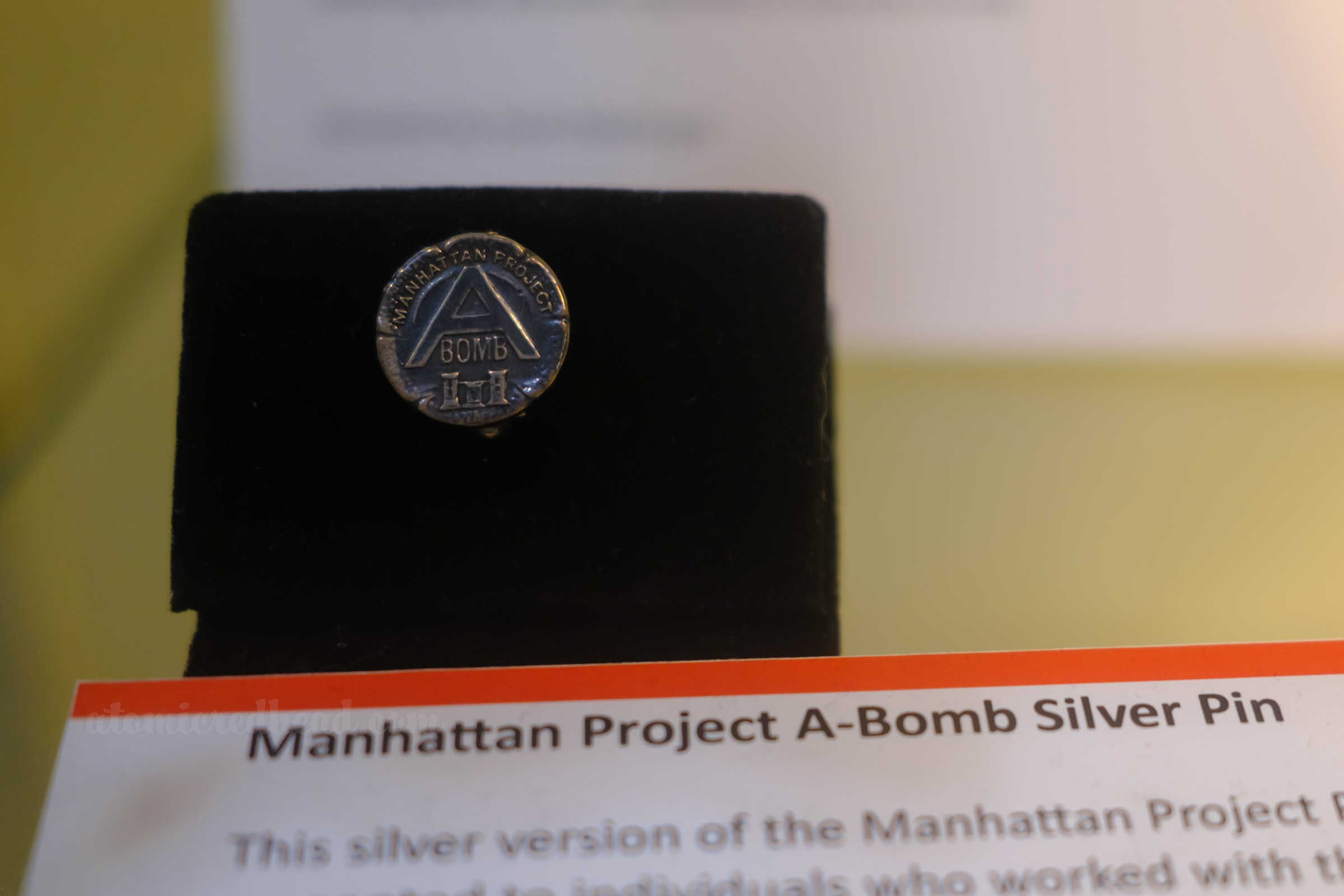 A small silver circular pin reads "Manhattan Project A Bomb" a gift to those who worked on the bomb.
