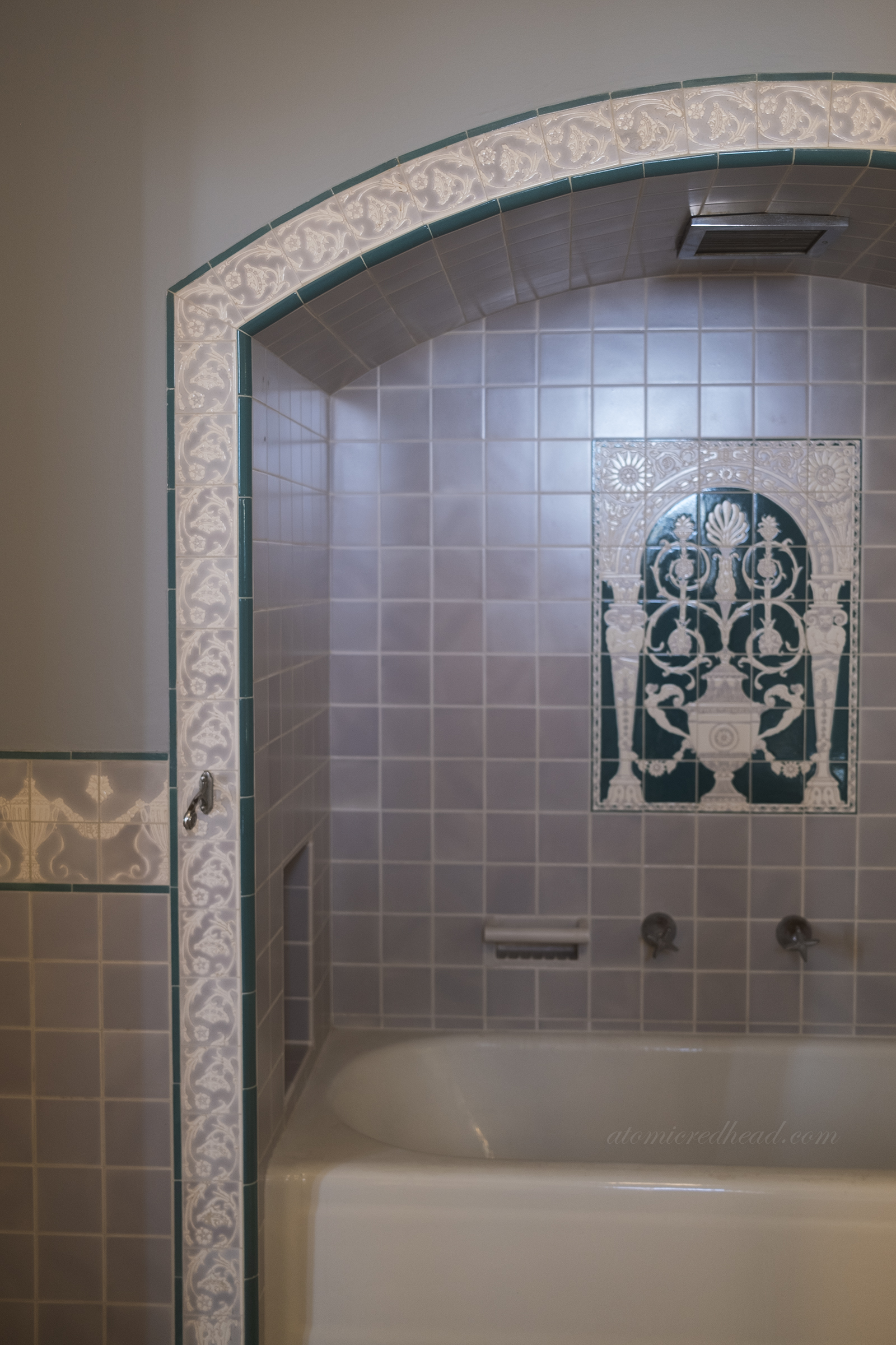 A dark teal and pale grey bathroom with arch over the tub, above the tub a tile mural with an ornate urn and column design.