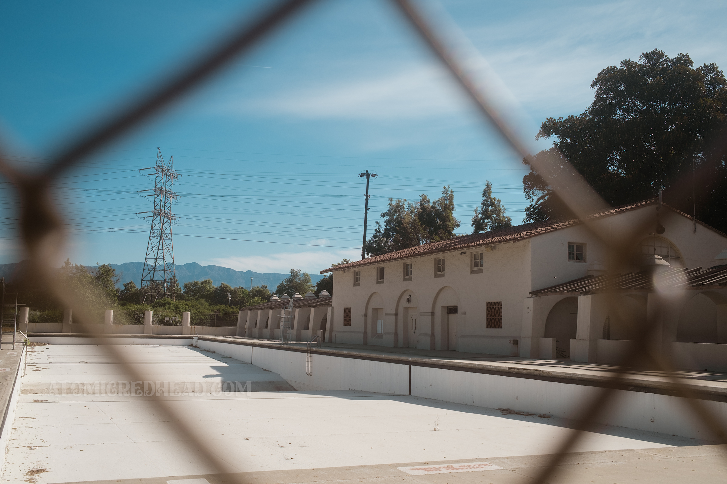 View of the long pool and its Spanish Revival style pool house as seen through a chainlink fence.