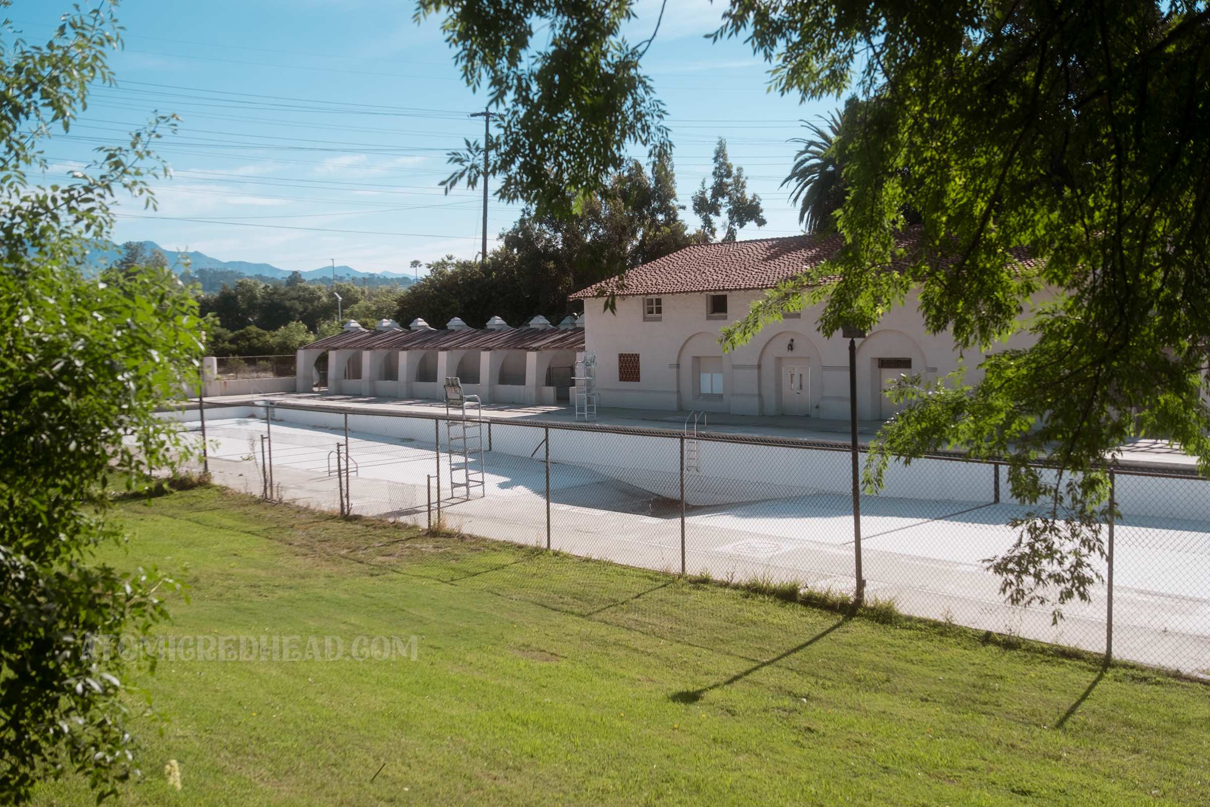View of the long pool and its Spanish Revival style pool house as seen through a chainlink fence.