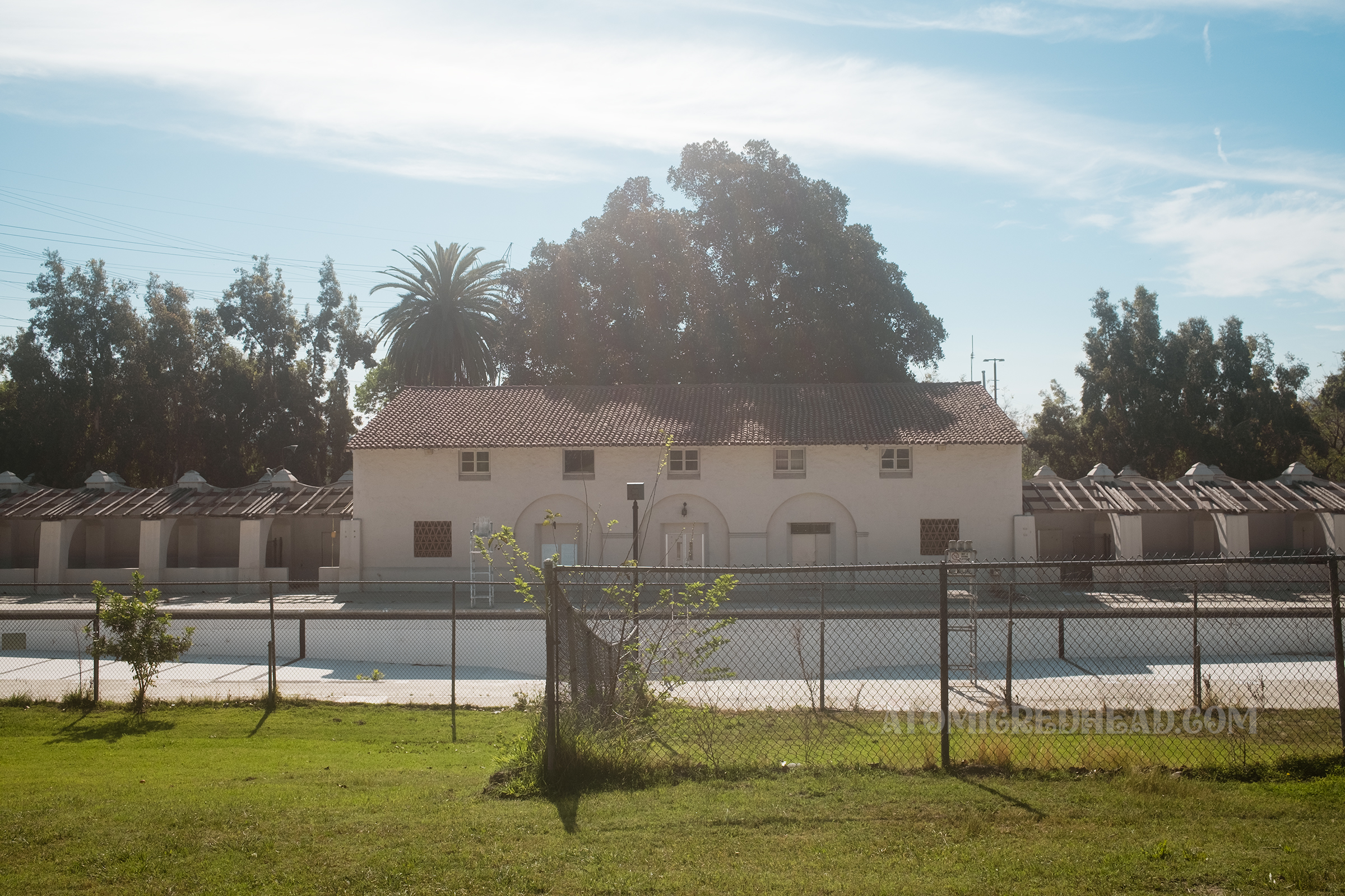 Straight on view as seen from a grassy hill next to the pool, a two story Spanish Revival style building with red tile roof sits in the middle, and is flanked by arched arcades on either side with the long empty pool in front.