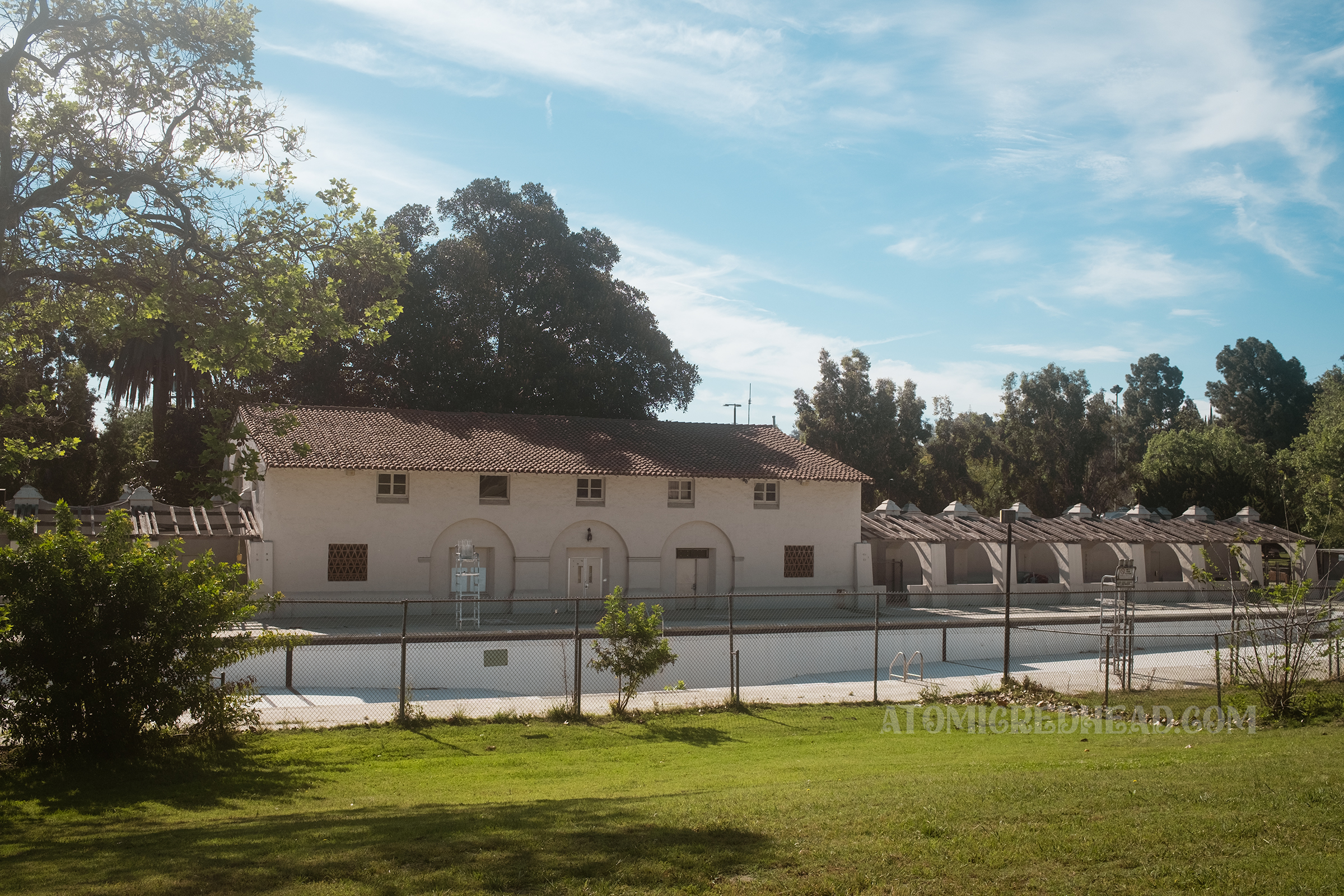 View of the long pool and its Spanish Revival style pool house as seen through a chainlink fence.