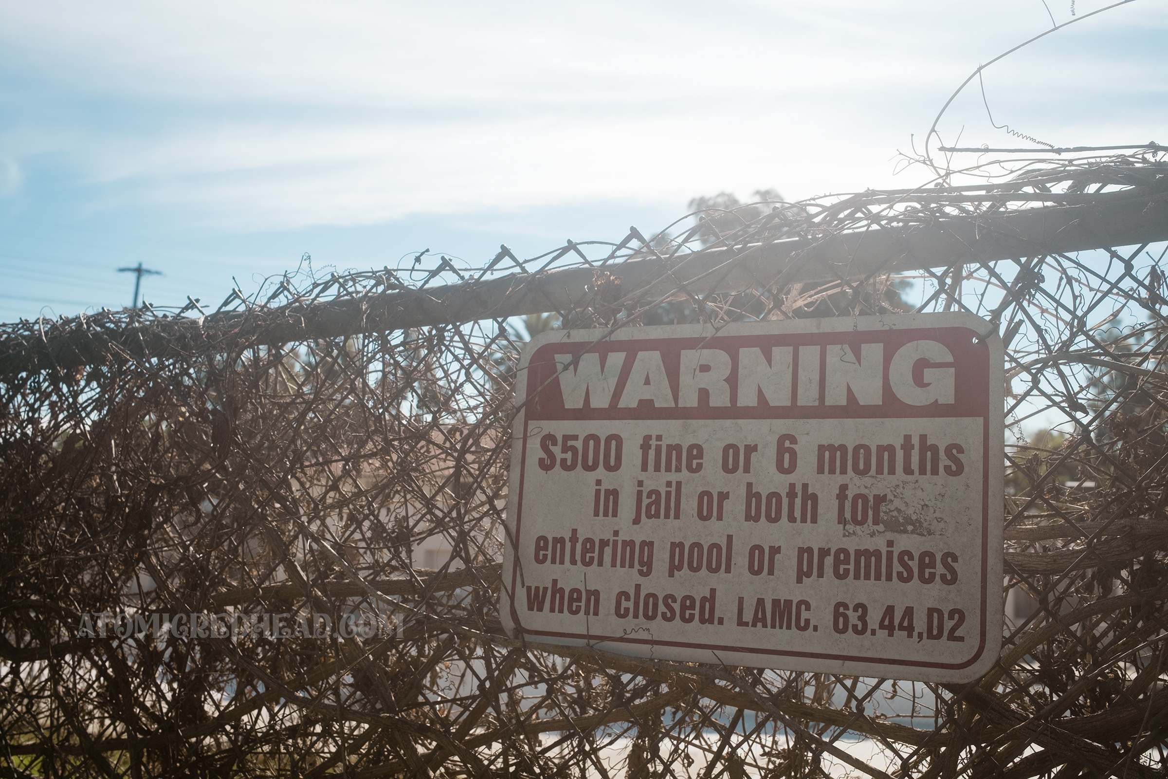 A red and white sign hangs on a chainlink fence reading "WARNING $500 fine or 6 months in jail or both for entering pool or premises when closed"