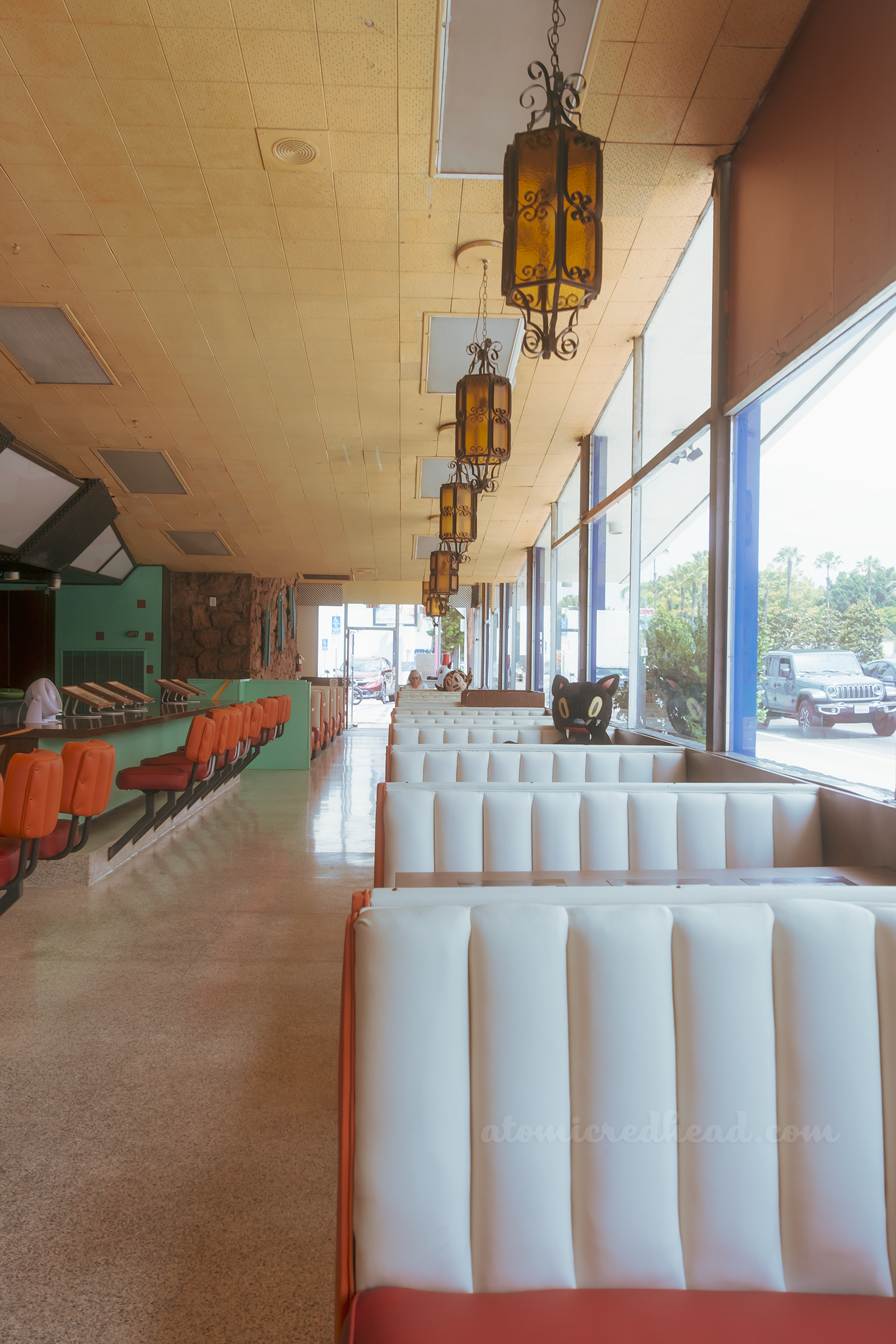 Rows of booths line windows on the right, with a large plush black cat sitting in one booth, to the left, orange bar stools line the counter.