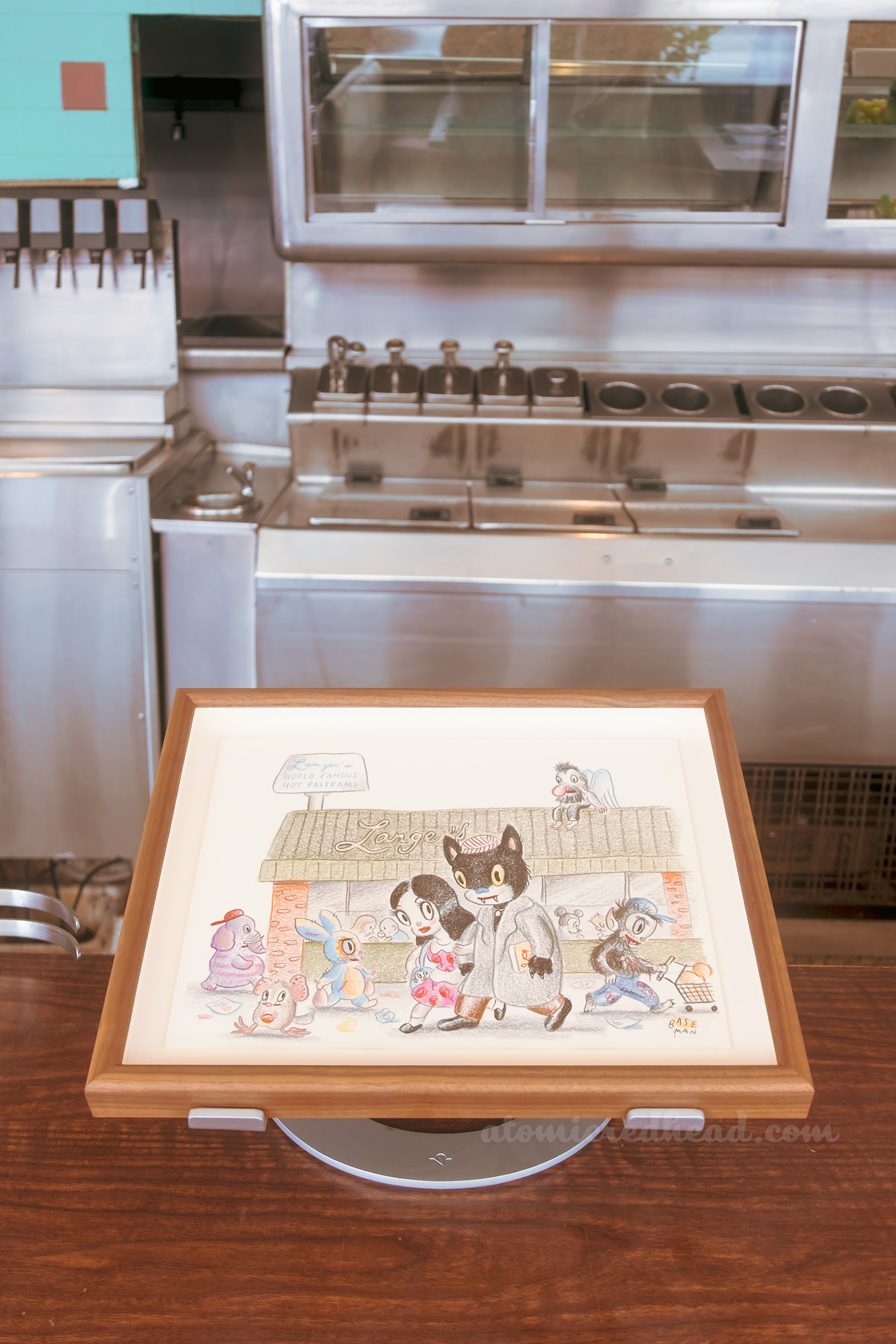 One of Baseman's works, this one features a black cat and a dark haired woman walking in front of Langer's Deli.