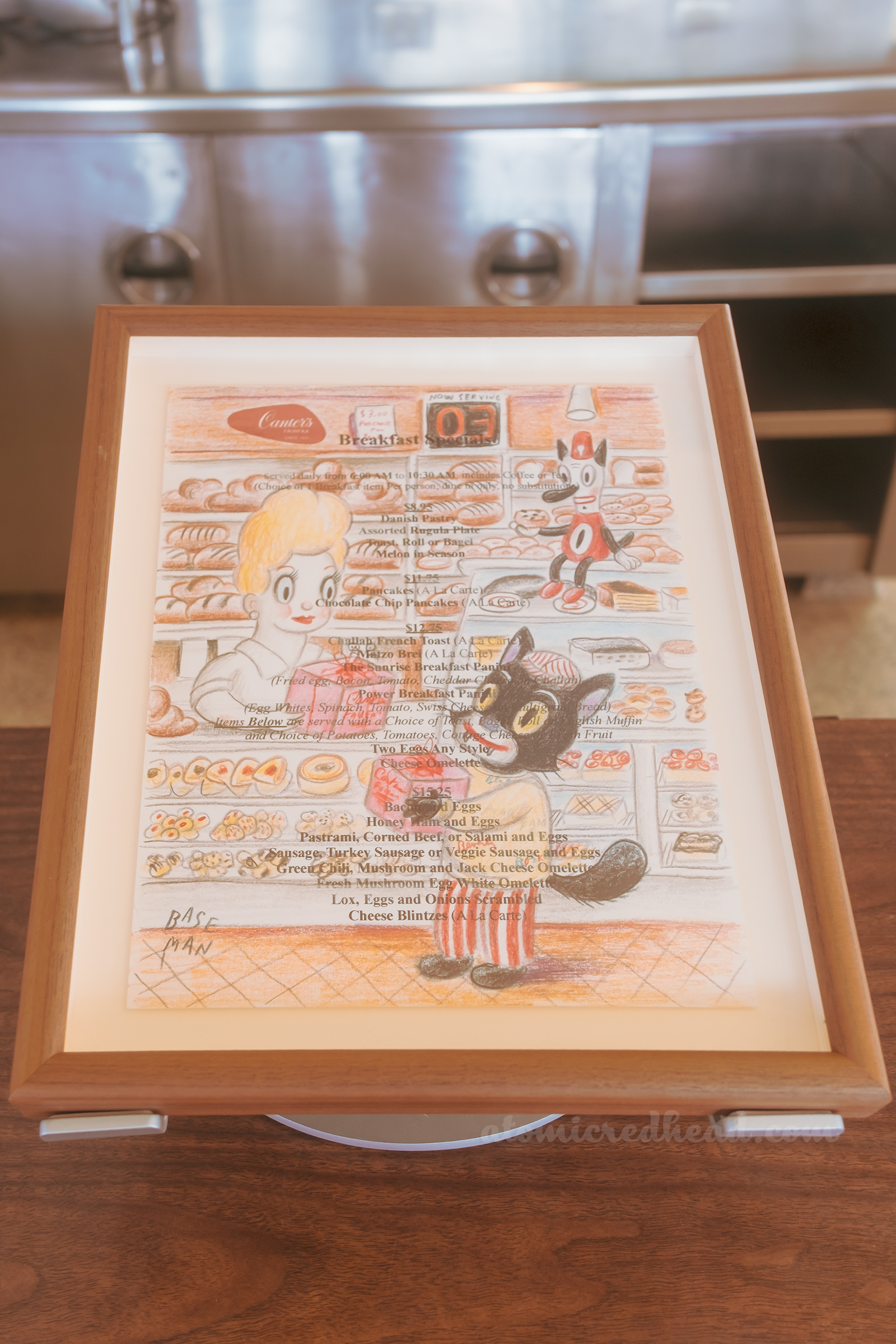 One of Baseman's works, this one features a black at holding a pink box at the register of Canter's Deli, a redhead with a beehive hairdo is behind the counter.