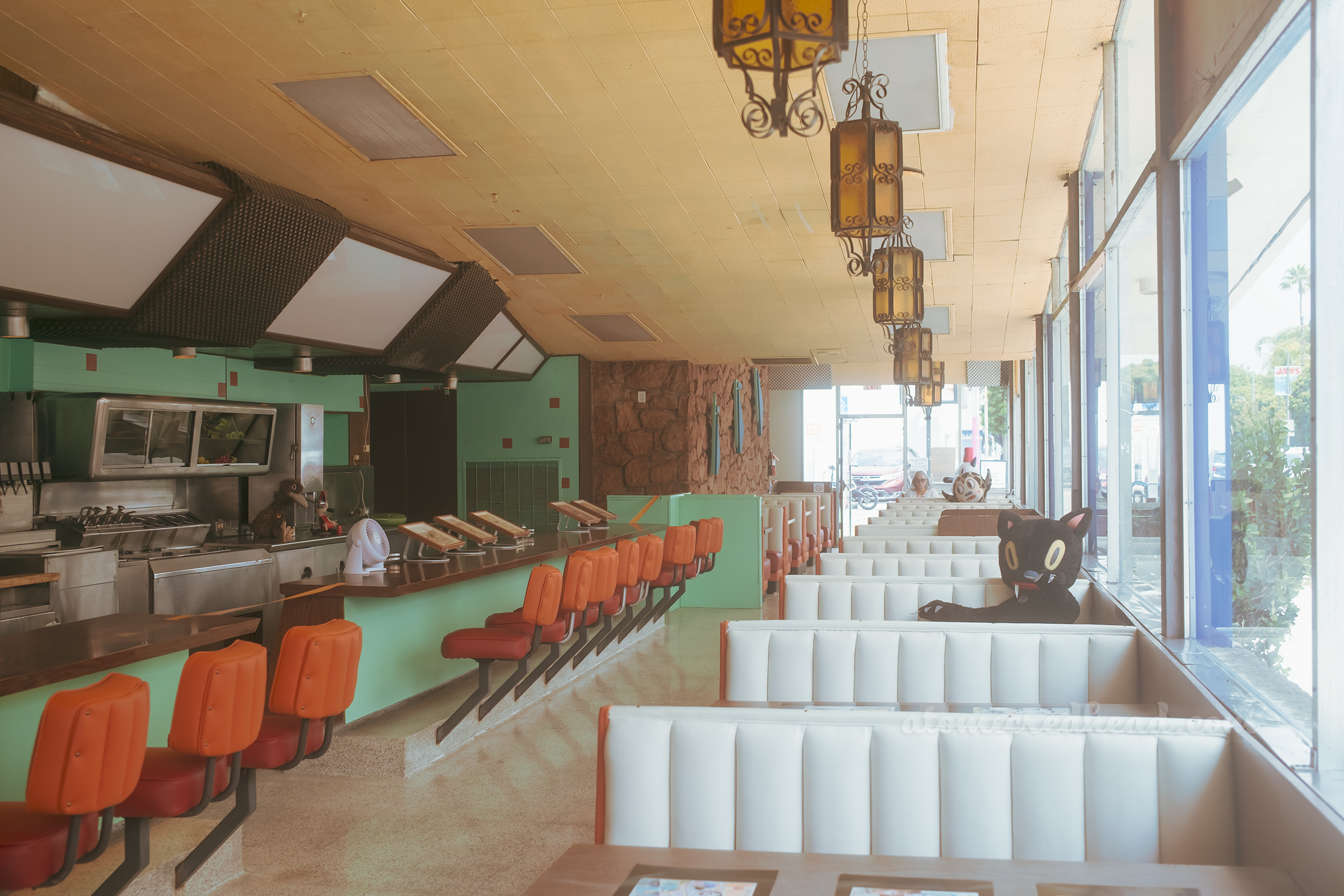 Rows of booths line windows on the right, with a large plush black cat sitting in one booth, to the left, orange bar stools line the counter.
