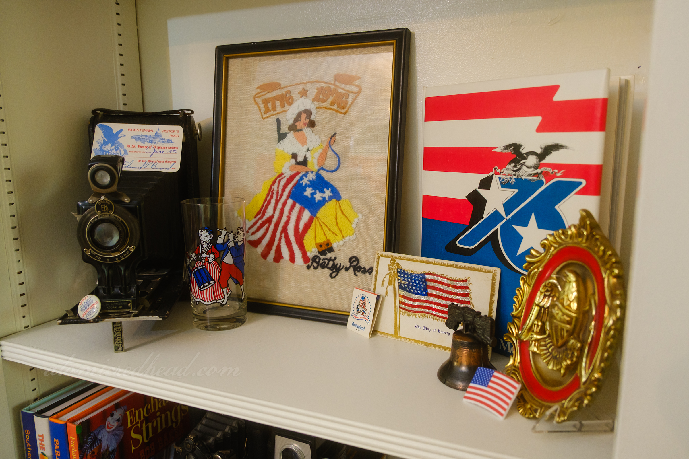 A shelf features an embroidered image of Betsy Ross sewing the American flag, a vintage book features a bald eagle and "76" in bold blue, and a vintage postcard of the American flag.