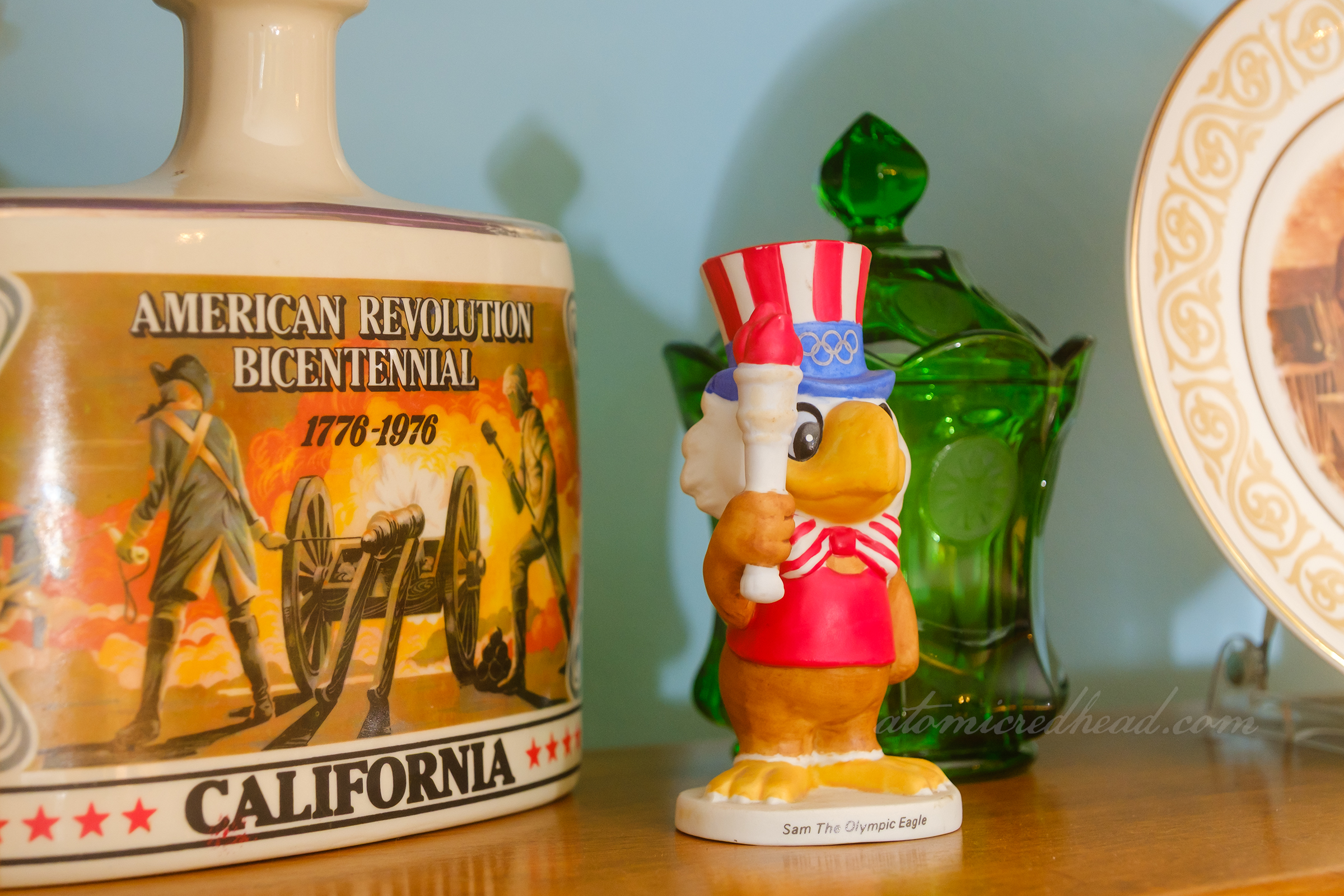Close-up of a ceramic figure of Sam the Eagle from the 1985 Olympic Games next to a bottle depicting a battle scene noting the arrival of California as a state.