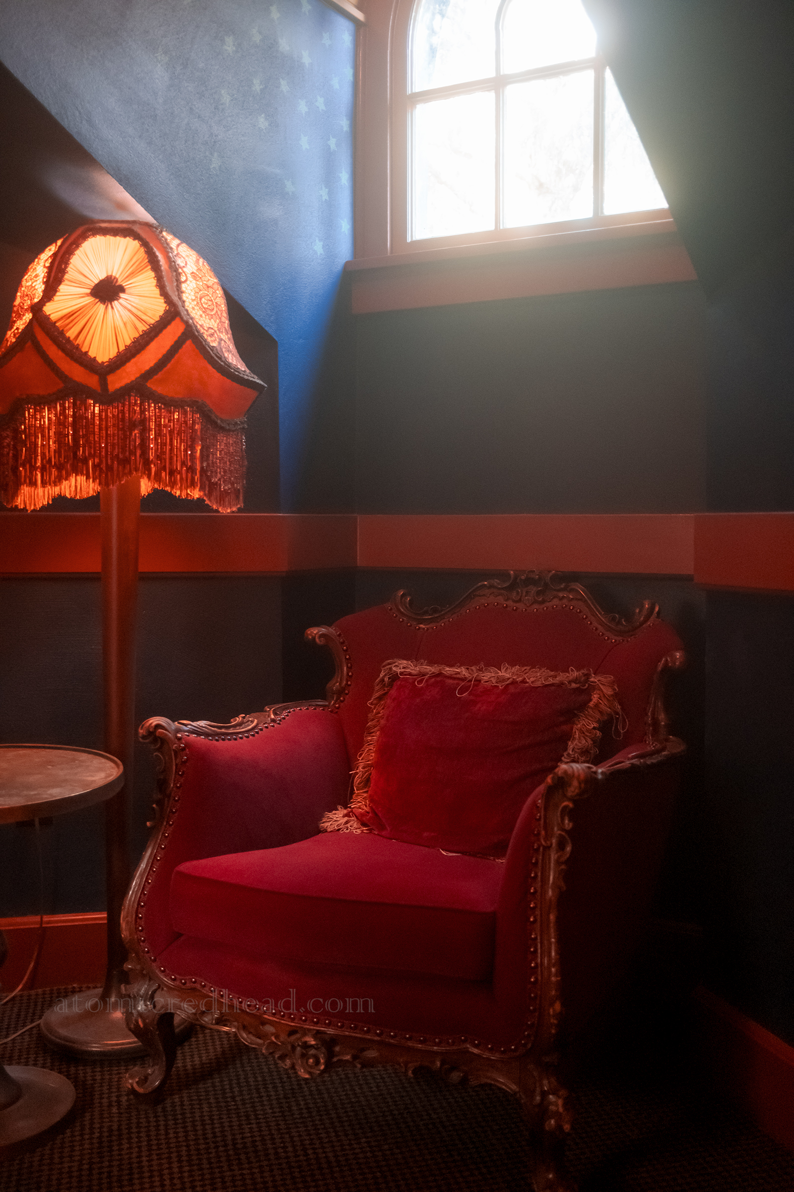 A red velvet chair sits next too a fringed lamp under a window and surrounded by rich dark blue walls.
