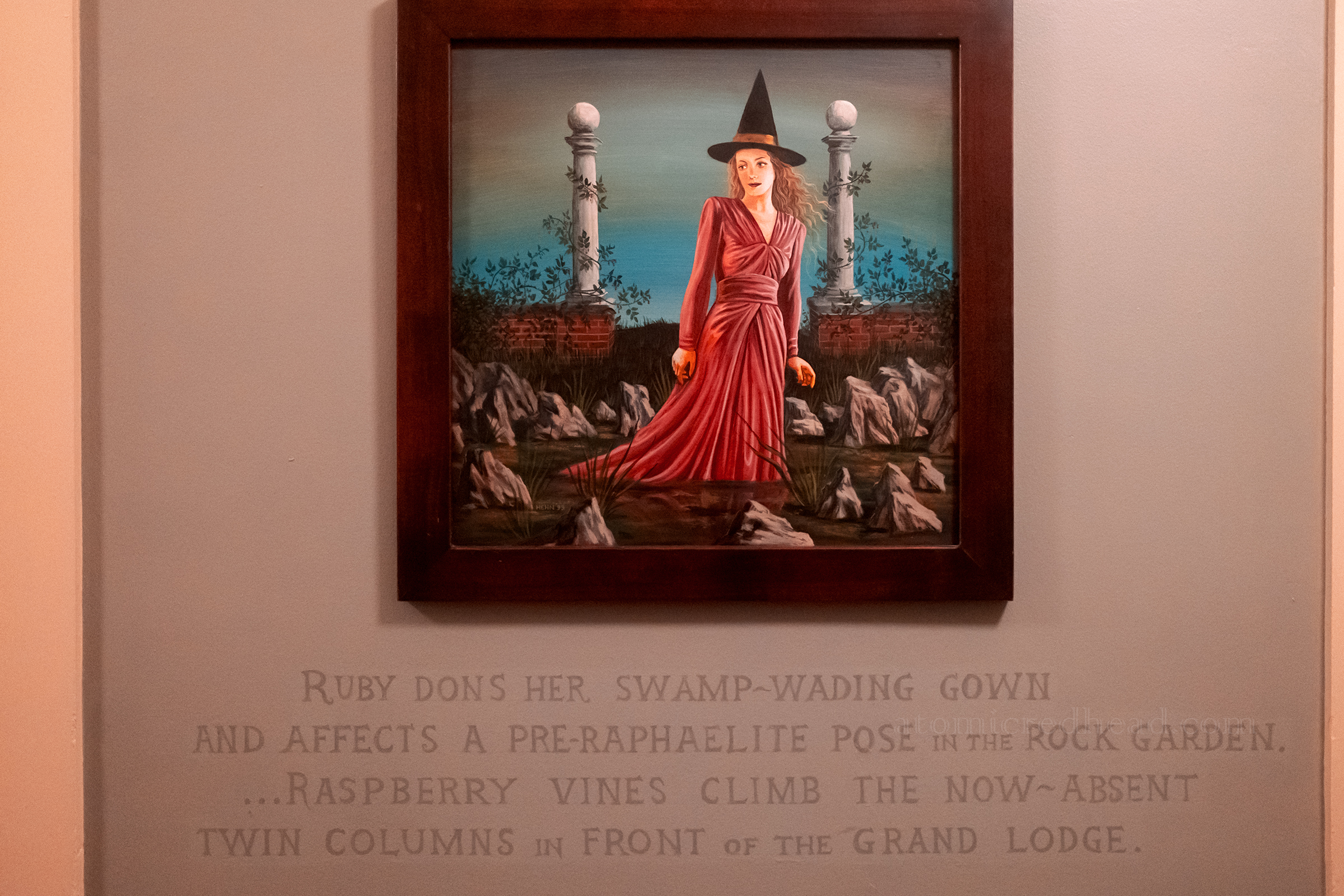 A painting of a woman in a red dress wearing a witch hat walks through water with rocks, below it reads "Ruby dons her swamp-wading gown and affects a pre-Raphaelite pose in the rock garden...Raspberry vines climb the now-absent twin columns in front of the Grand Lodge."