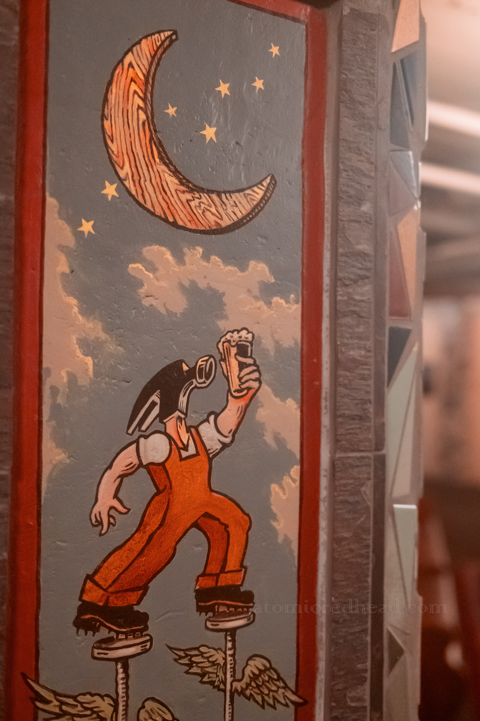 A painting of the McMenamin character Hammerhead, a man with a hammer for a head, wearing orange overalls holds a pint of beer under a crescent moon.